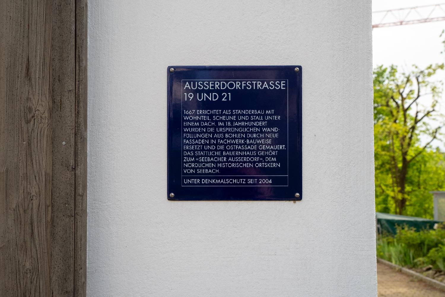 A blue plate on a wall, with the following German text: "Ausserdorfstrasse 19 und 21 - 1667 errichtet als Ständerbau mit Wohnteil, Scheune und Stall unter einem Dach. Im 18. Jahrhundert wurden die ursprünglichen Wandfüllungen aus Bohlen durch neue Fassaden in Fachwerk-Bauweise ersetzt und die Ostfassade gemauert. Das stattliche Bauernhaus gehört zum «Seebacher Ausserdorf», dem nördlichen historischen Ortskern von Seebach. Unter Denkmalschutz seit 2004".