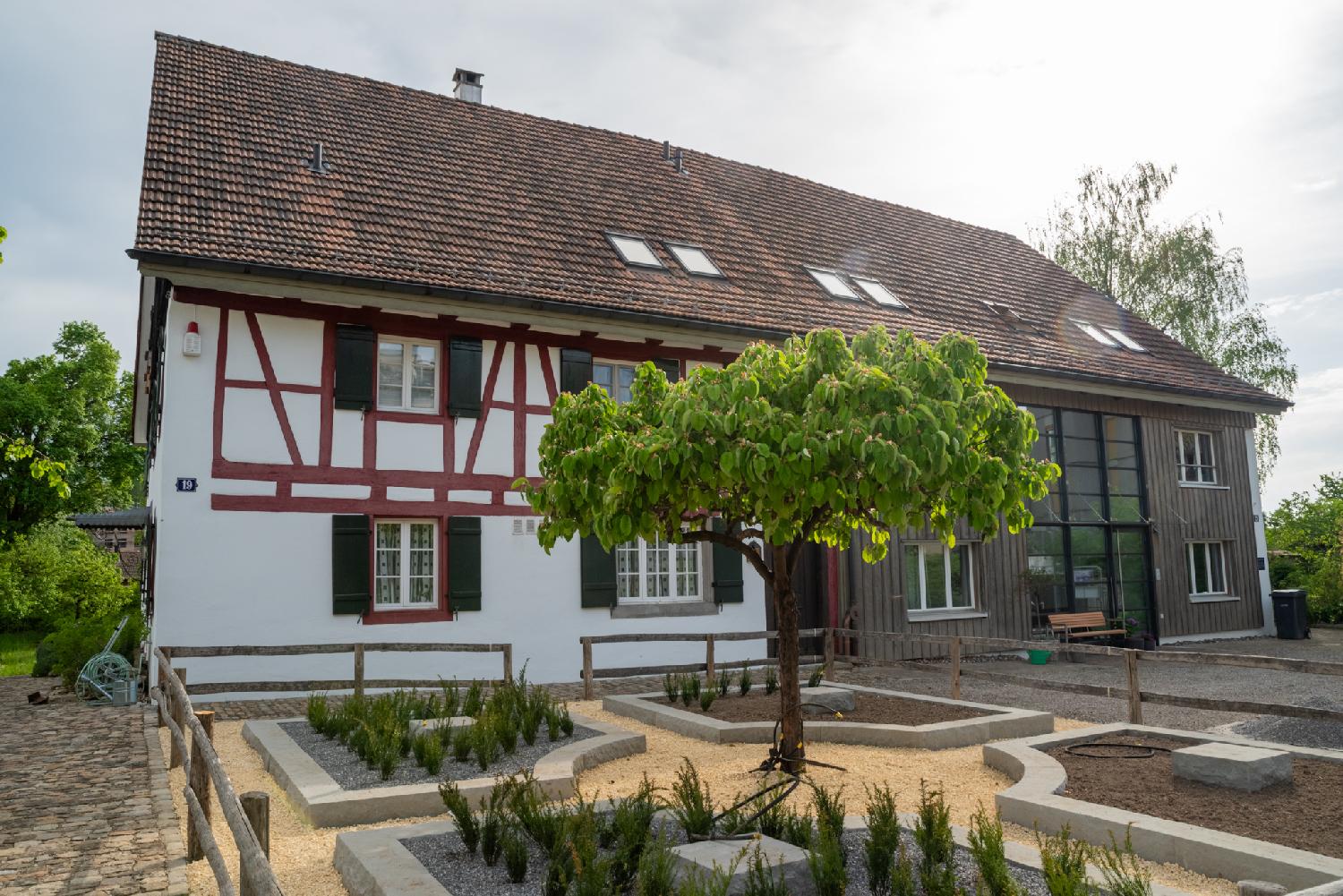 A building made of two parts: a half-timbered white and red building, and a wooden part on the right, under the same tiled roof. In the foreground, a small yard with a tree and a few planters.