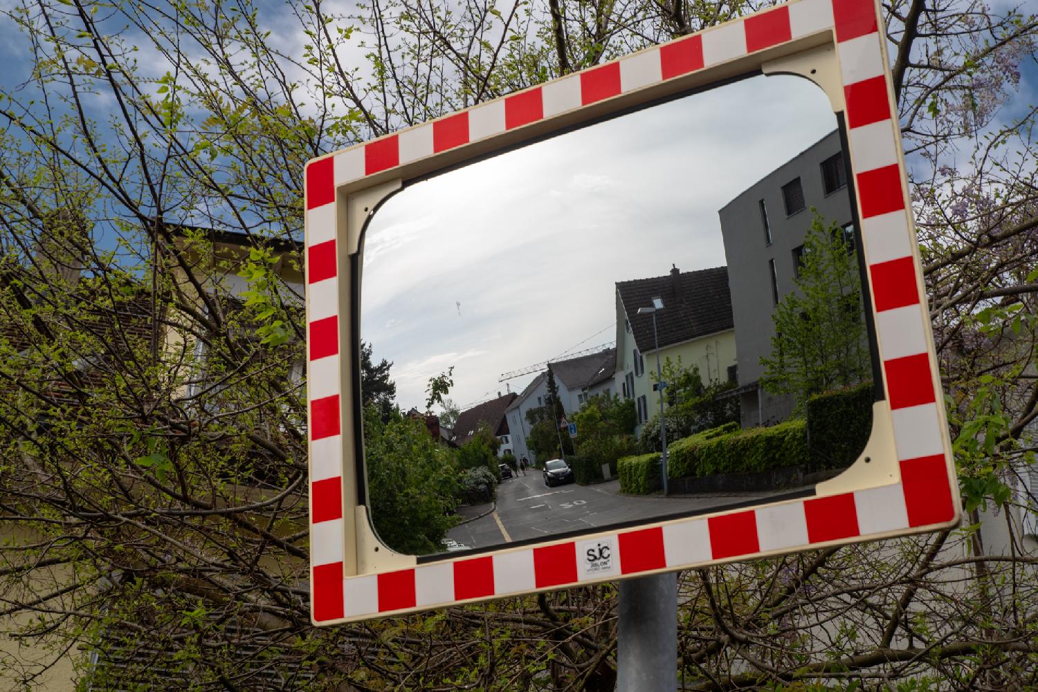 A traffic mirror with red and white border, showing a residential street with 2-3-story building behind hedges