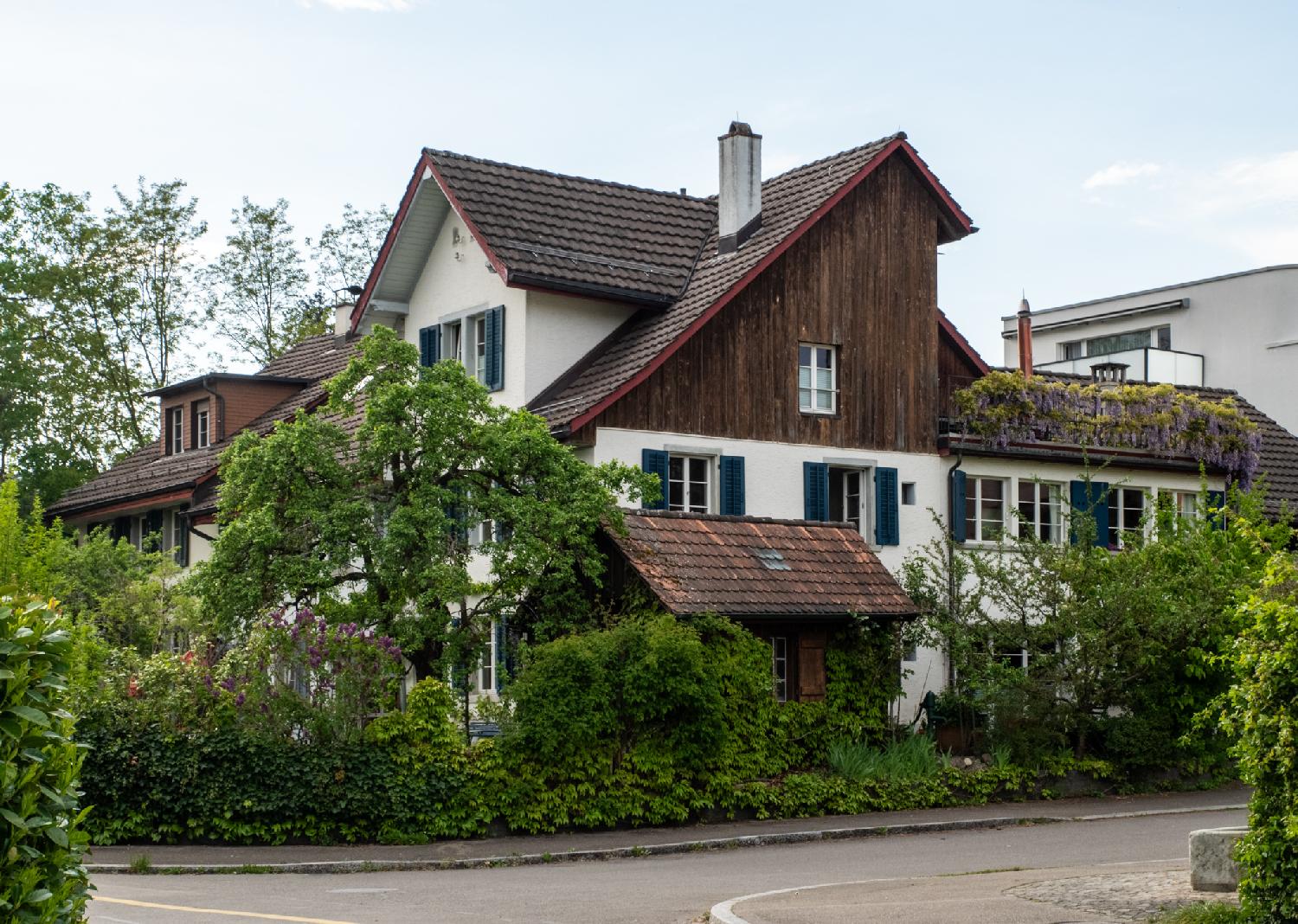 A residential construction with a lot of angles and façades, including a wooden façade, behind hedges and with a lush garden.