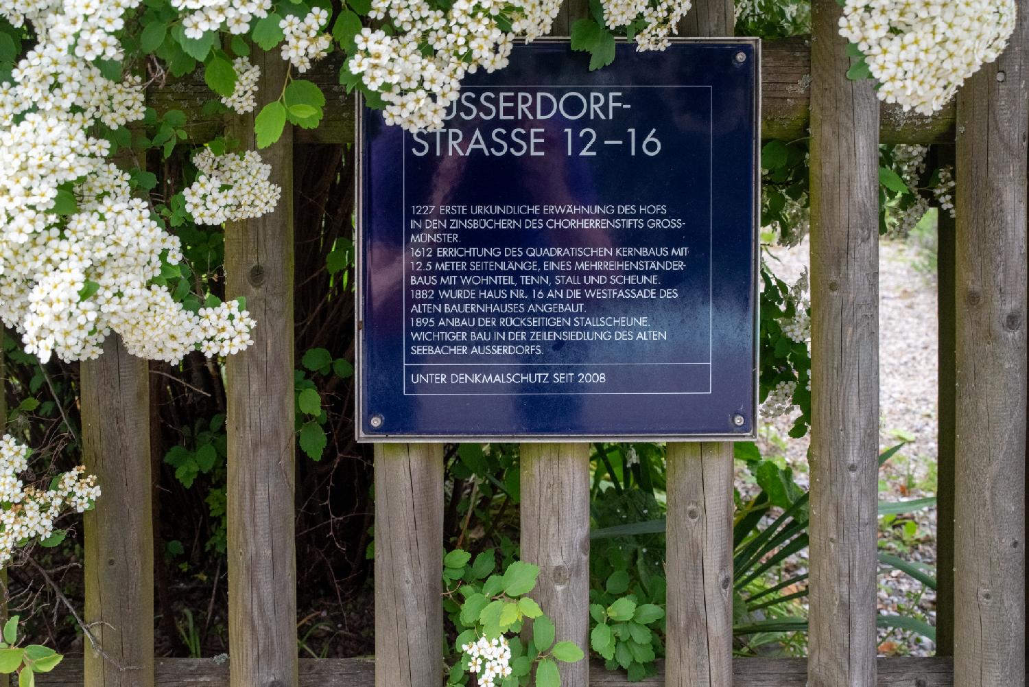 A blue plate no a wooden fence, surrounded by white flowers, with a text in German: "Ausserdorfstrasse 12-16 -  1227 erste Urkundliche Erwähnung des Hopfs in den Zinsbüchern des Chorherrenstifts Grossmünster.  1612 Errichtung des Quadratischen Kernbaus mit 12.5 Meter Seitenlänge, eines Mehreihenständerbaus mit Wohnteil, Tenn, Stall und Scheune. 1882 wurde Haus Nr. 16 an die Westfassade des alten Bauernhauses angebaut. 1895 Anbau der Rückseitigen Stallscheune. Wichtiger Bau in der Zeilensiedlung des Alten Seebacher Ausserdorfs. Unter Denkmalschutz seit 2008."