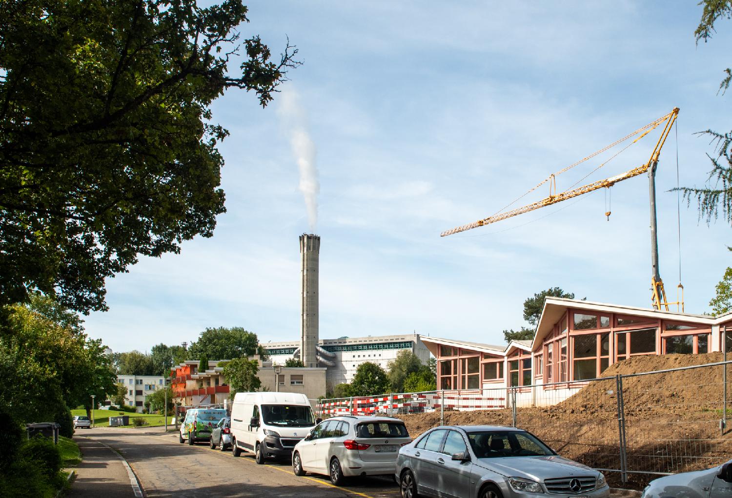 A narrow street with a row of cars parked on the right, a construction site, and a smoking chimney
