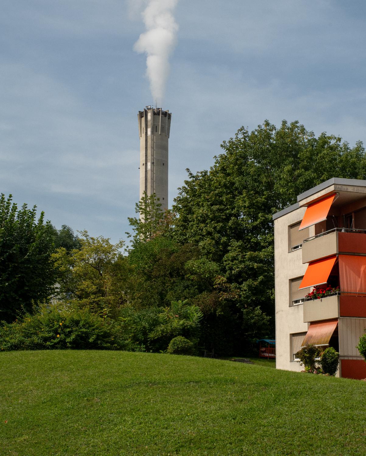 A smoking grey chimney behind trees behind a building with orange sunscreens, and a lawn in the foreground.