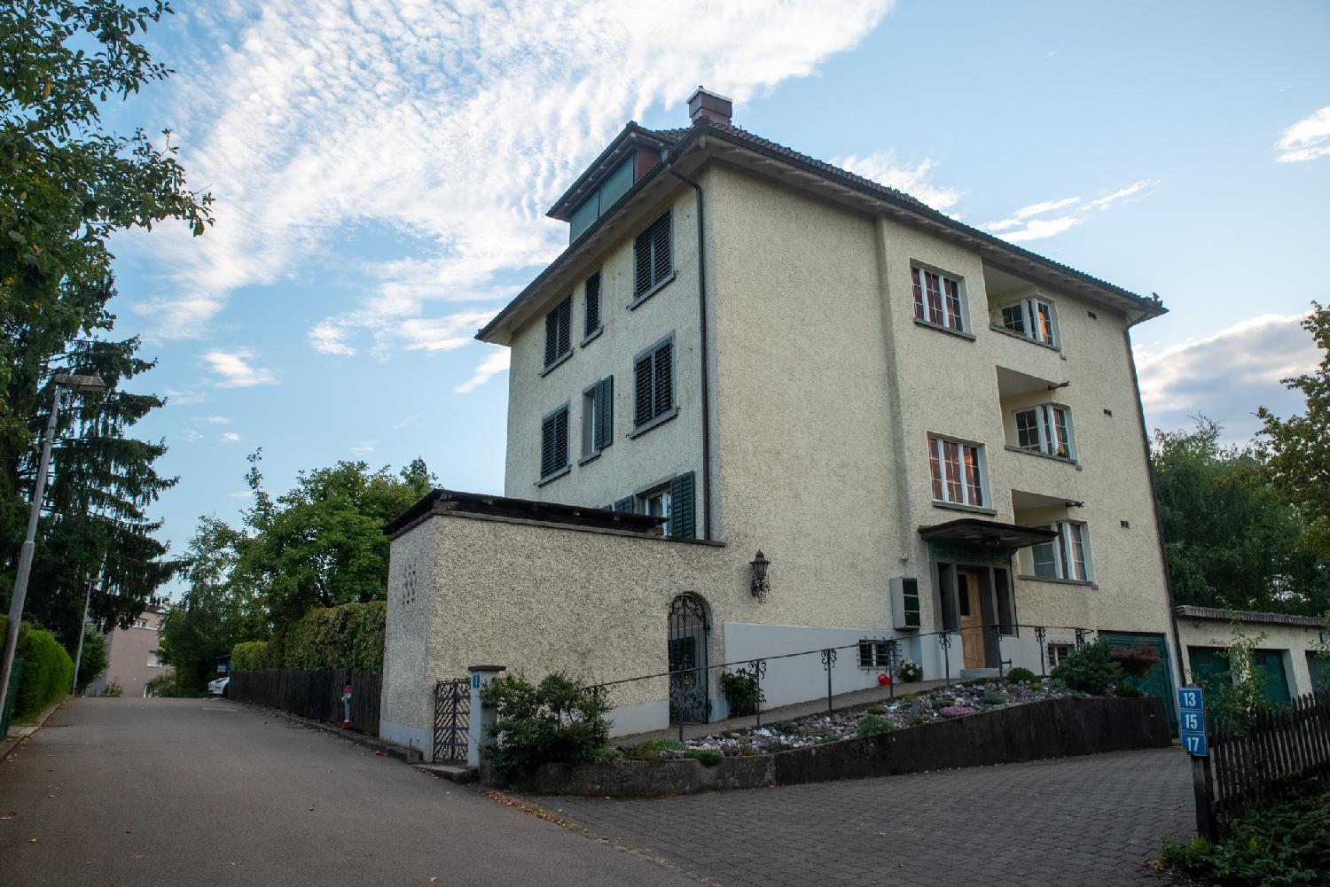 A narrow street with a 4-story residential yellow building with green blinds in the foreground. The building has a rounded door on its side.