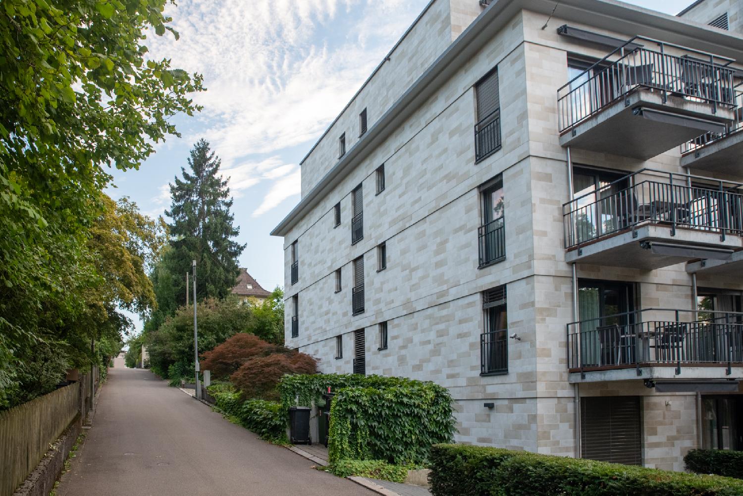 A narrow street with a wooden fence and trees on the left, and a modern 5-story beige residential building with visible stones and large balconies on the right