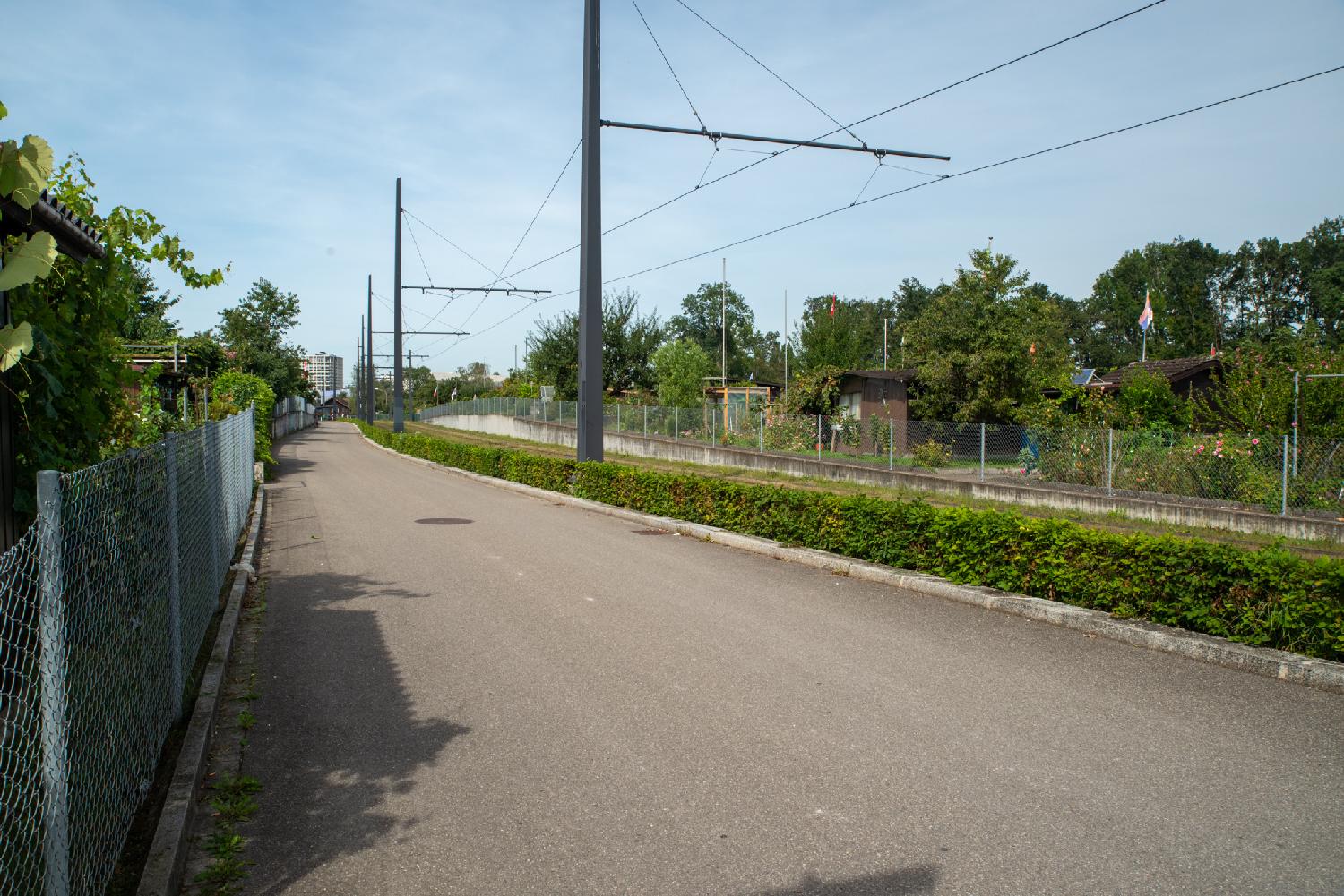 A street with no markings; on the left, a metallic fence with gardens behind; on the right, a tram railway with electric lines.