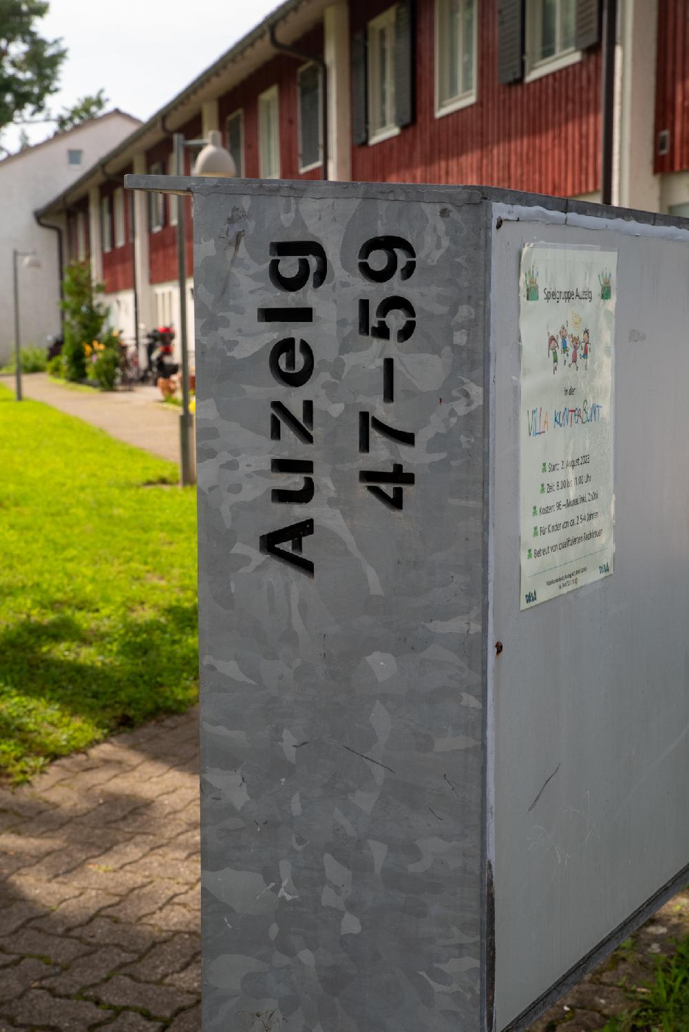 A grey set of mailbox with "Auzelg 47-59" cut in the metal, in front a row of of 2-story red houses