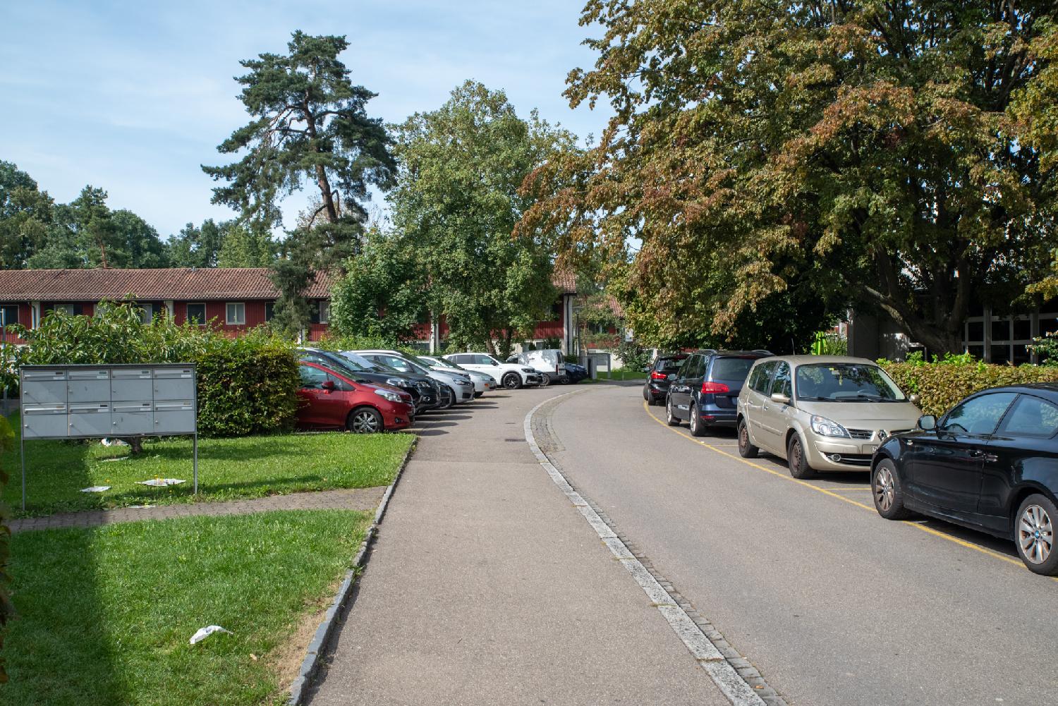 A narrow street with cars on the right parked in front of a hedge below a tree, and another parking lot on the left in front of 2-story rows of red houses