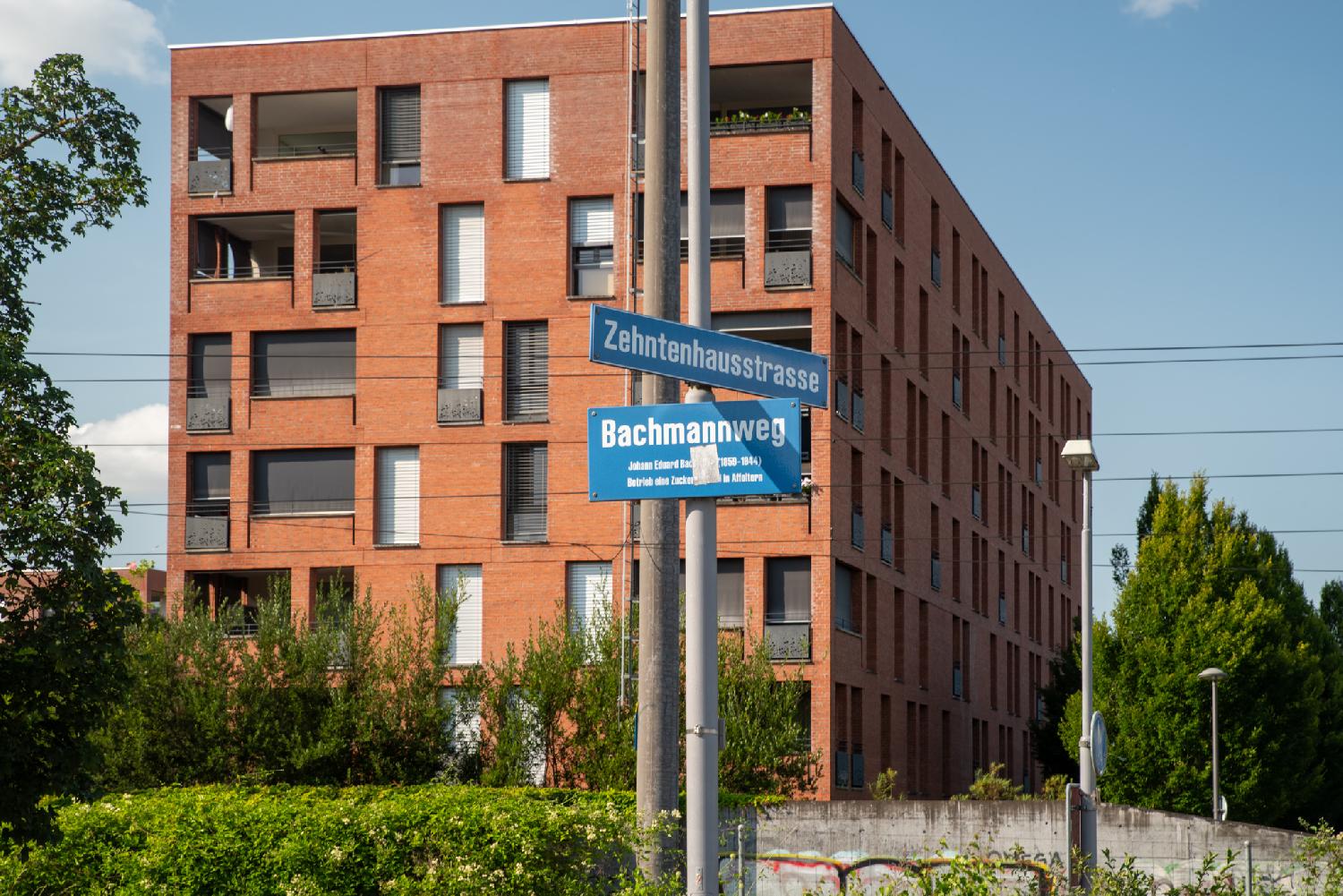 A metallic pole with two blue street signs for Zehntenhausstrasse and Bachmannweg in front of a red/orange residential building.