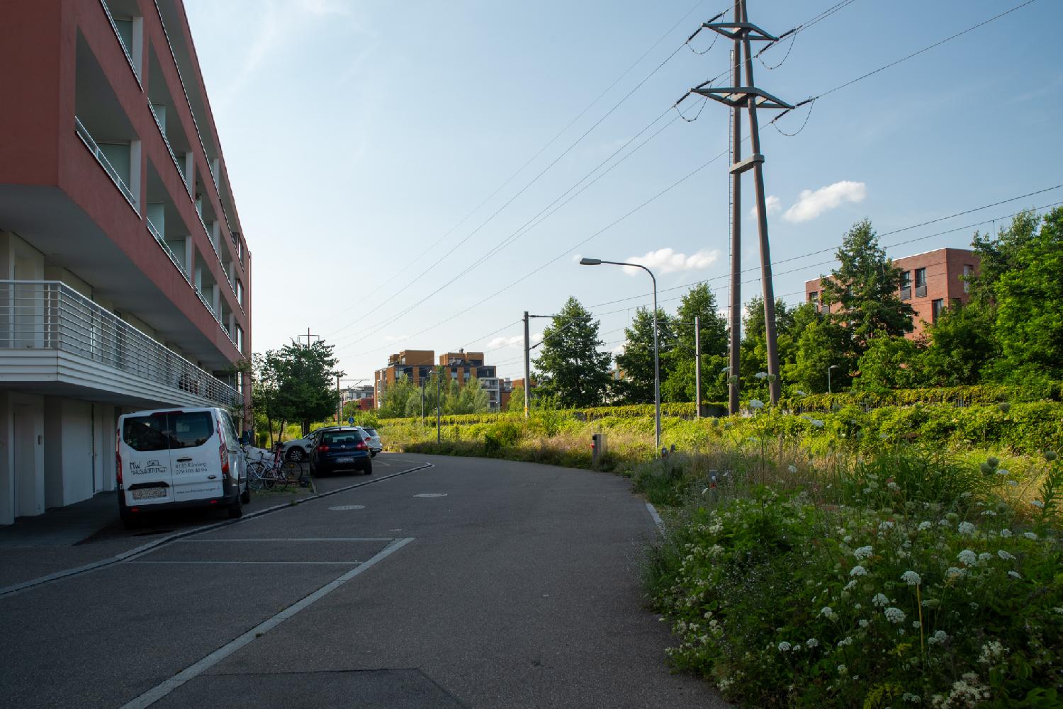 A street with a red building and cars on the left side and a train track on the right side. Other red buildings are visible in the background and on the other side of the tracks.
