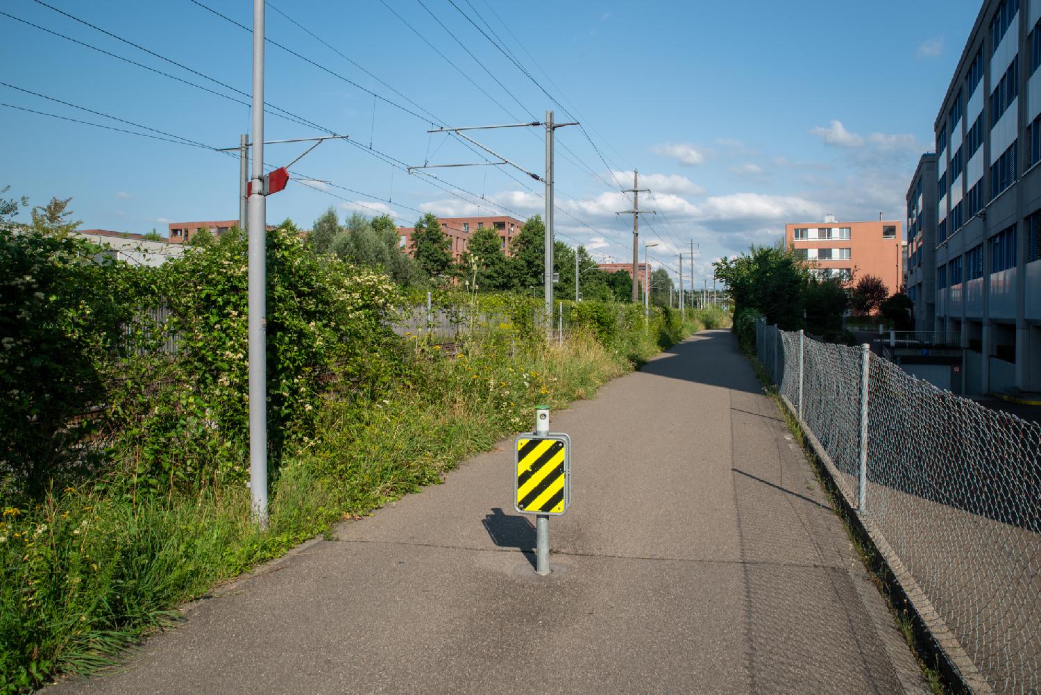 A narrow road with a yellow and black zebra sign in the middle of it, buildings on the right and train tracks on the left