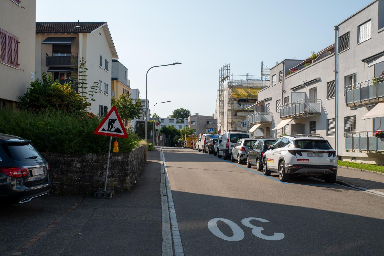 A residential street with buildings on both sides, a row of cars on the left, a "30" painted on the road, and building work in the background