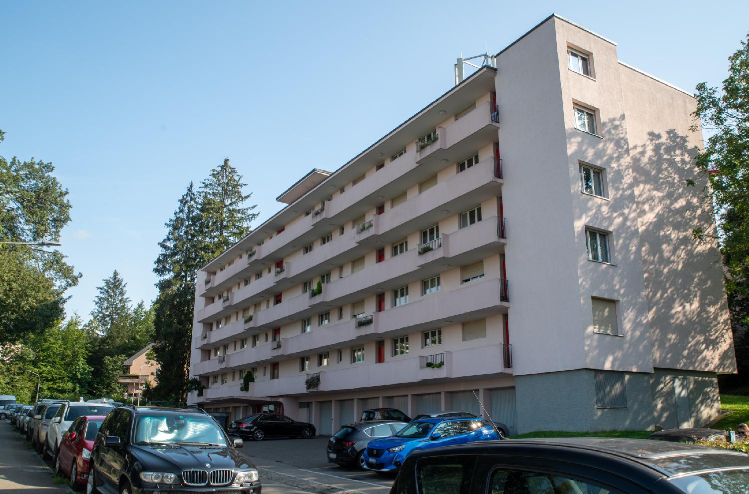 A six-story light pink residential building with garage doors on the ground floors and a lot of cars parked on the street in front of it