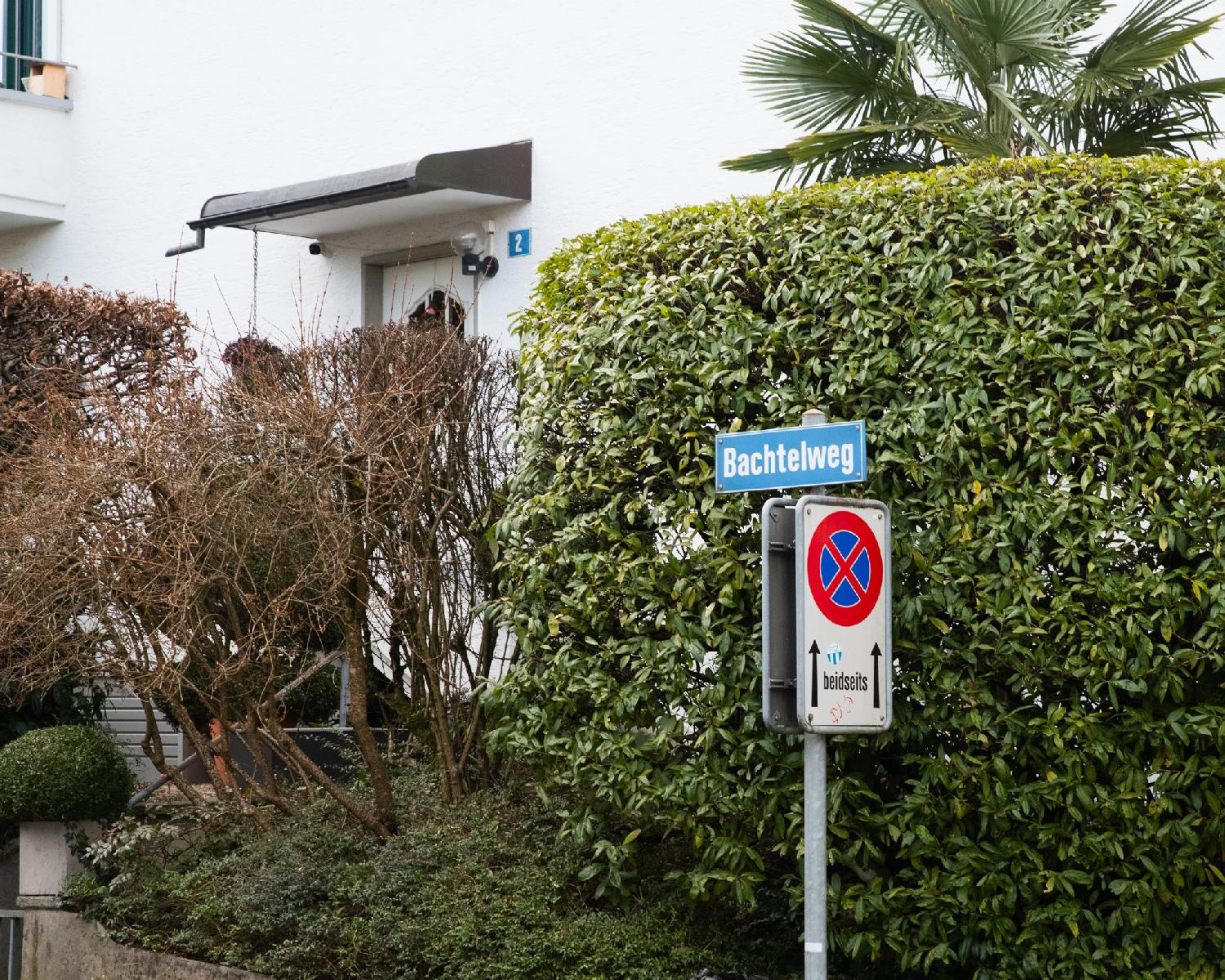 A blue Bachtelweg street sign, on top of a no-parking sign, in front of a green hedge next to a white house numbered 2.