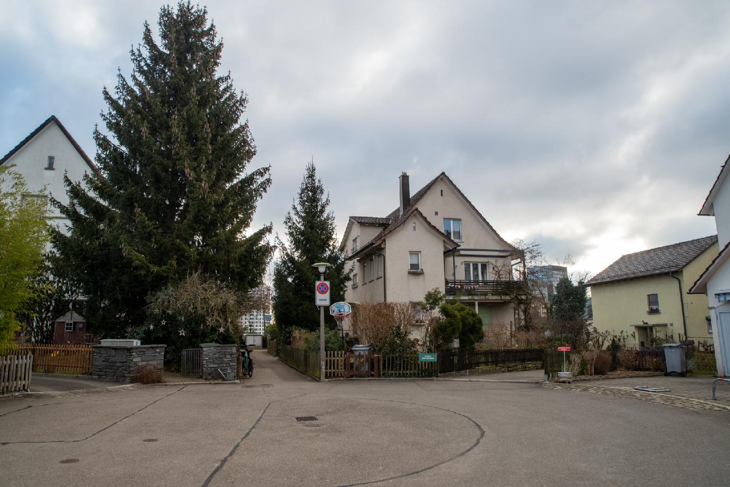 A round square at the end of a residential street, with a narrow pedestrian alley opposite the photograph. There are a few homes, a large conifer tree with stars attached at the bottom of it, a basketball hoop and a no parking sign.