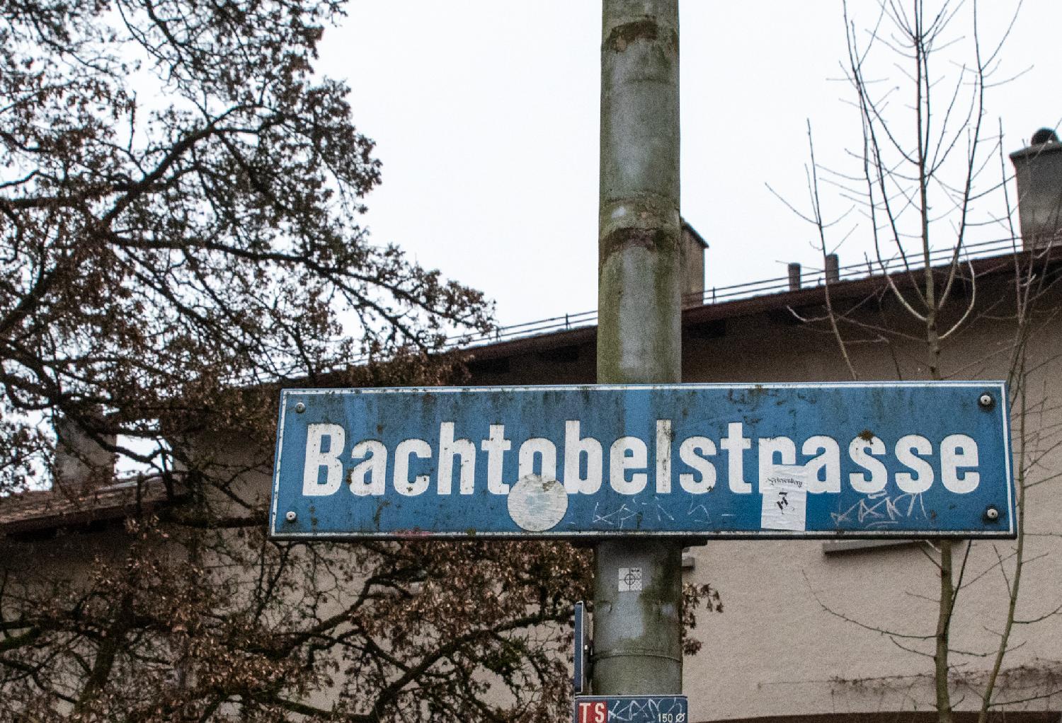 A grey pole in front of a residential building and trees, with a dirty "Bachtobelstrasse" blue sign attached to it