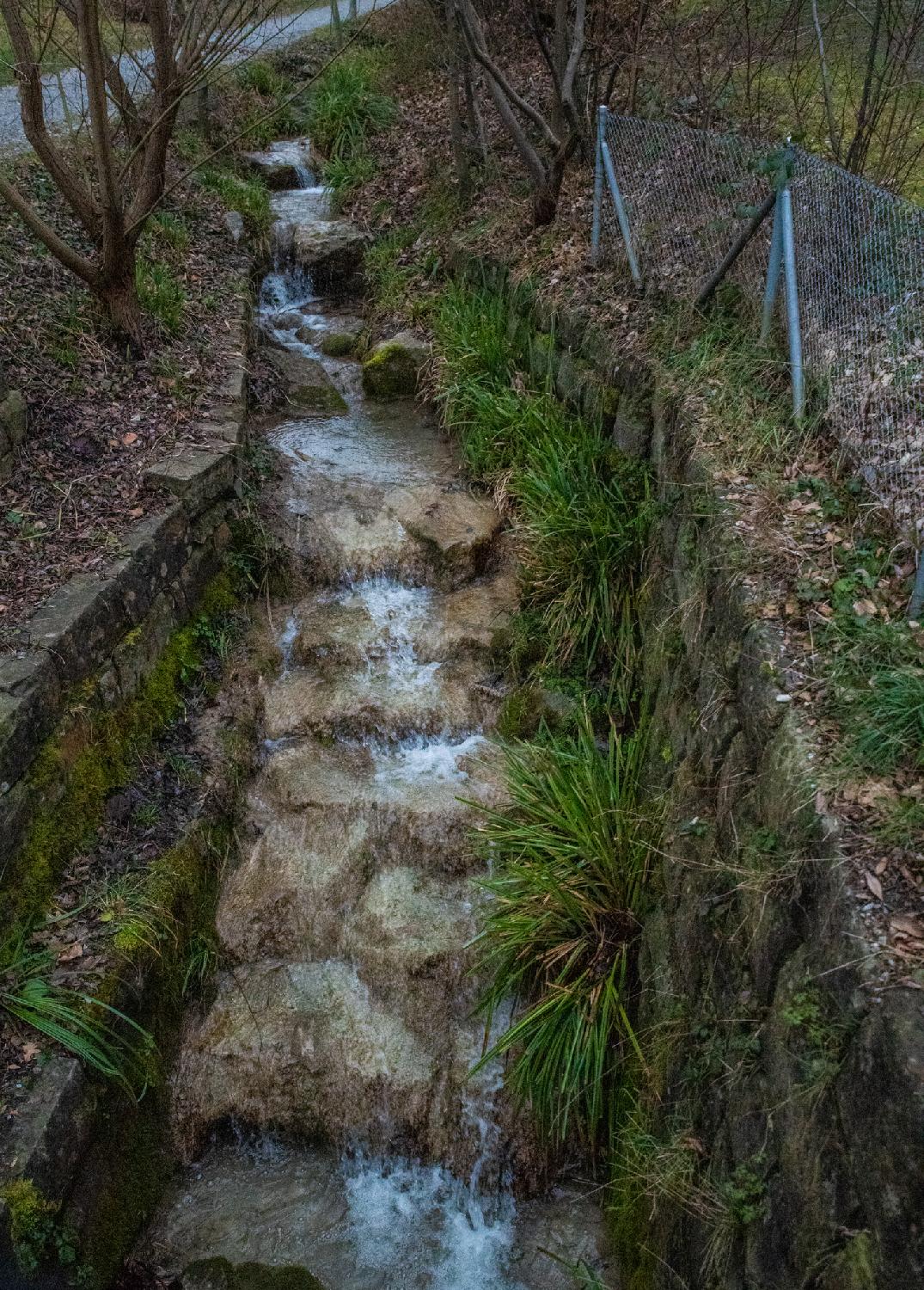 A small water stream falling on light brown steps, with vegetation-covered walls on each side