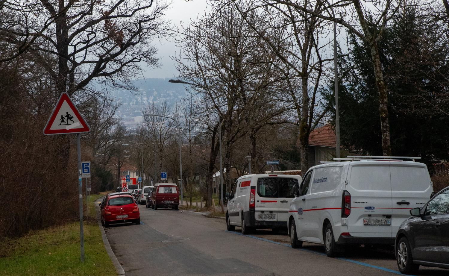 A road going downhill, with cars parked on each side under trees without leaves, and a hill with buildings visible in the hazy background