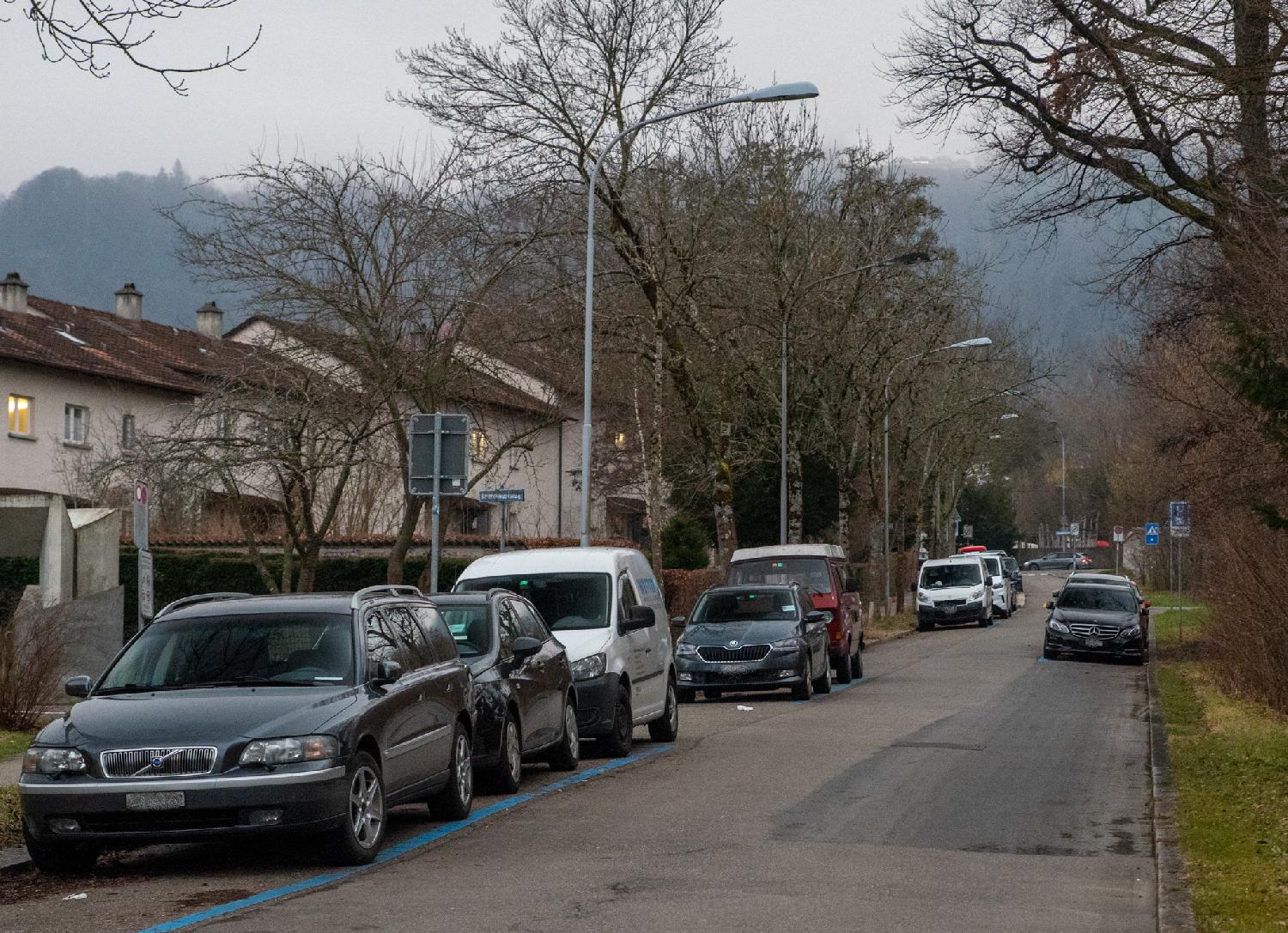 A one-way street with cars on each side, low residential buildings on the left side, trees on both sides, and a hazy, woody hill in the background