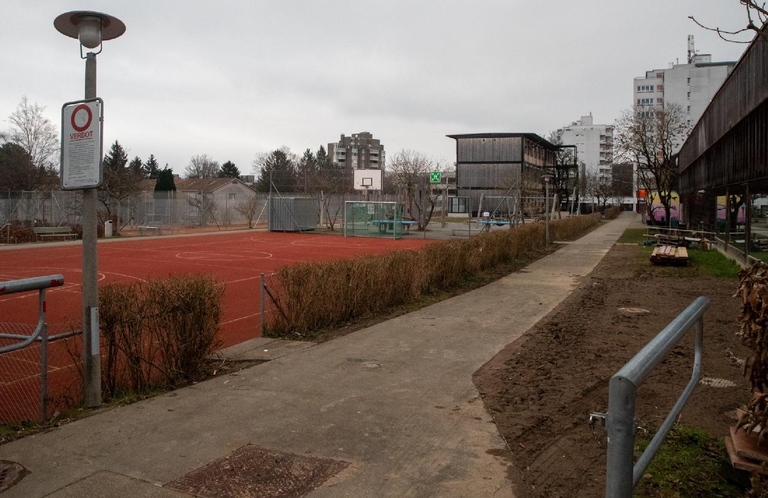 A red/orange tartan basketball court with a pedestrian path on its right side; a few buildings including a wooden prefab school building in the background