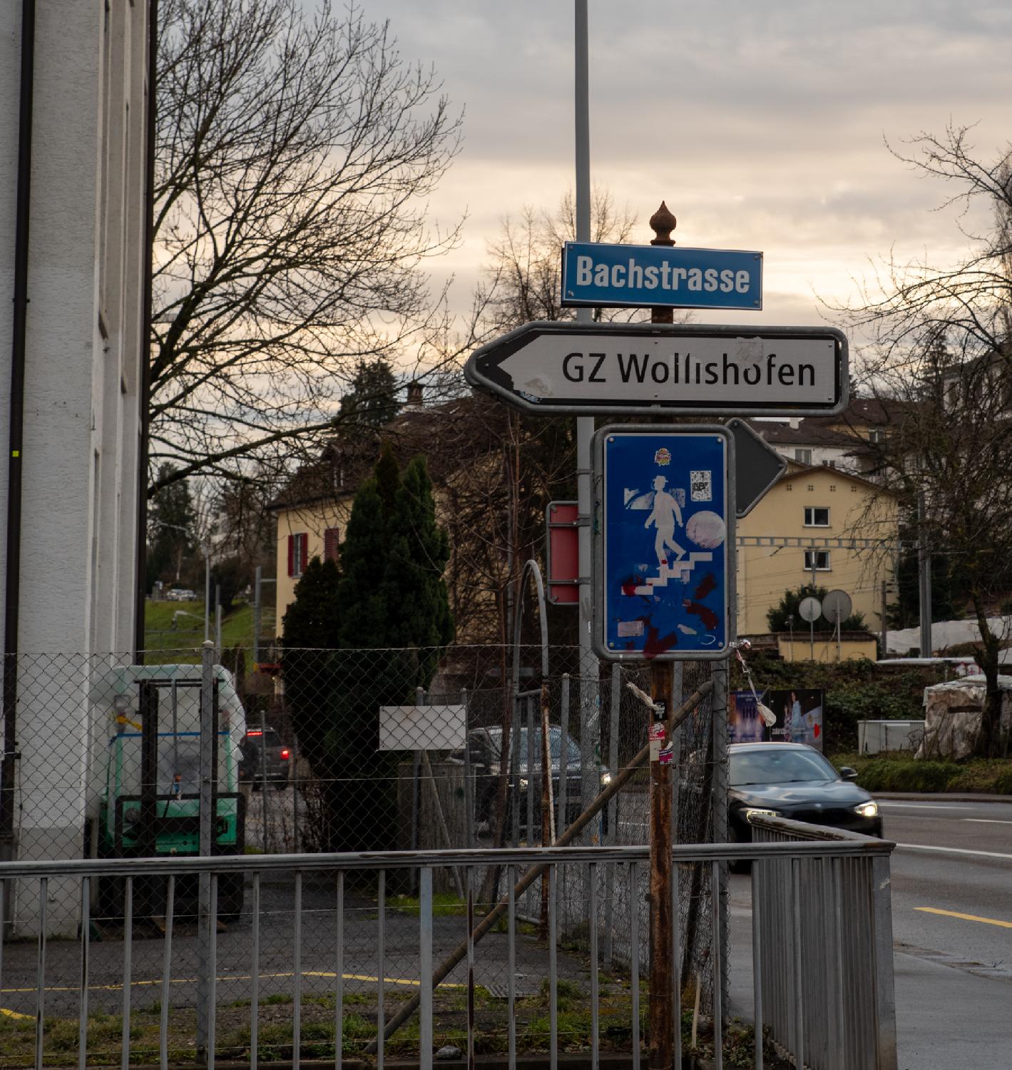 Corner of a street with a sign post containing a blue Bachstrasse street sign, a direction to GZ Wollishofen, and a pedestrian undeground passage sign with a lot of old stickers on it. In the background, a few yellow buildings and a road with traffic.