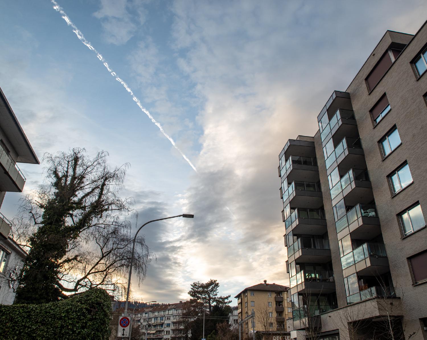 Buildings on the right side of the street, with windows reflecting a blue sky. On the right side, a tree covered with ivy behind a hedge. The sky is showing twilight colors and a plane trail splits the sky in two.