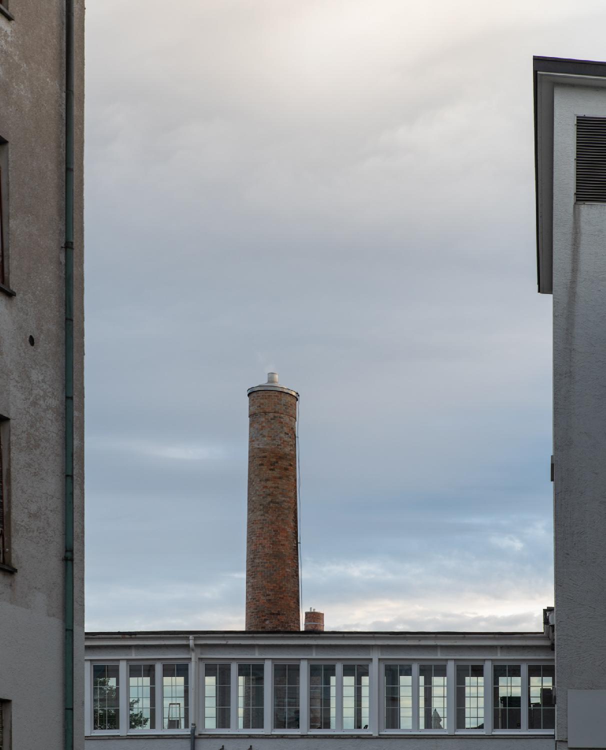 A brick chimney between two higher buildings, behind a covered bridge with a lot of glass windows