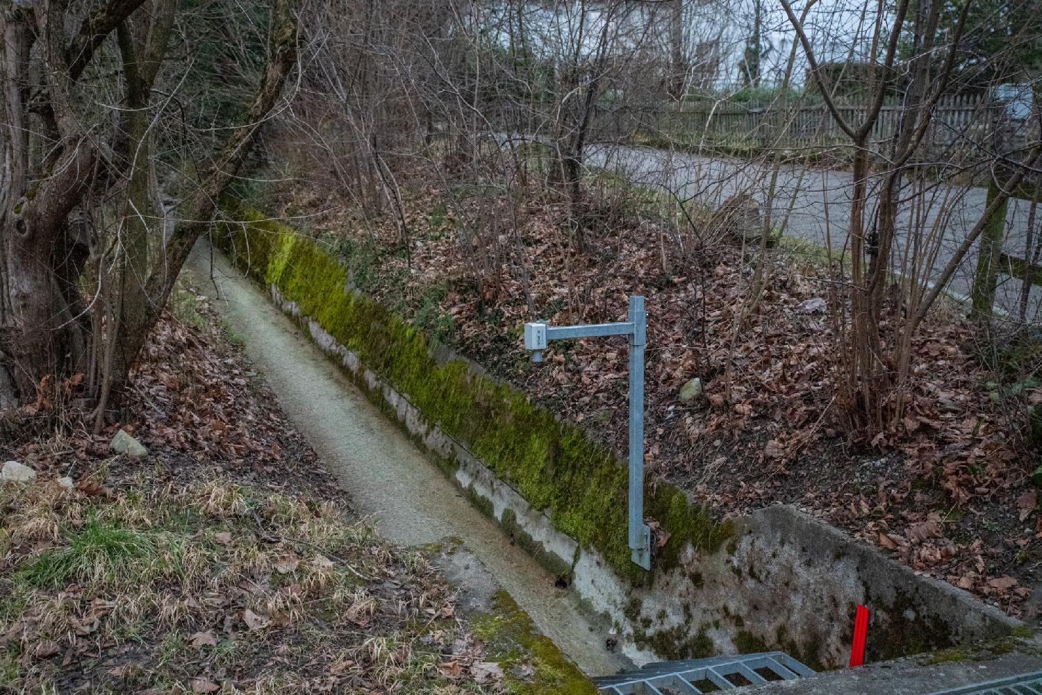 A small water stream in a concrete canal, with a camera attached to a metallic pole pointing at it