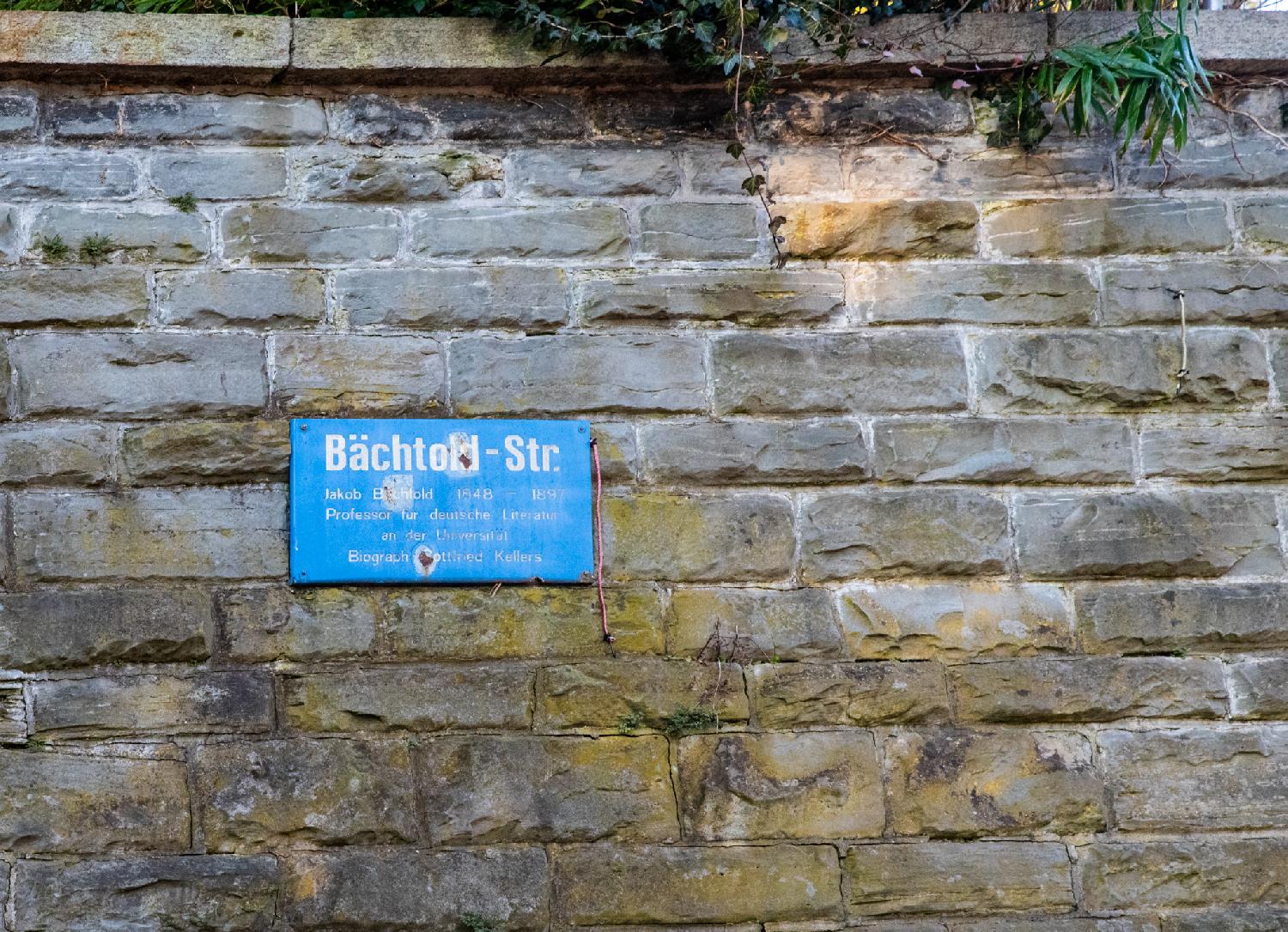 A stone wall with a blue street sign that reads "Bächtold-Str. - Jakob Bächtlod 1848 - 1897 - Professor für deutsche Literatur and der Universität - Biograph Gottfried Kellers"
