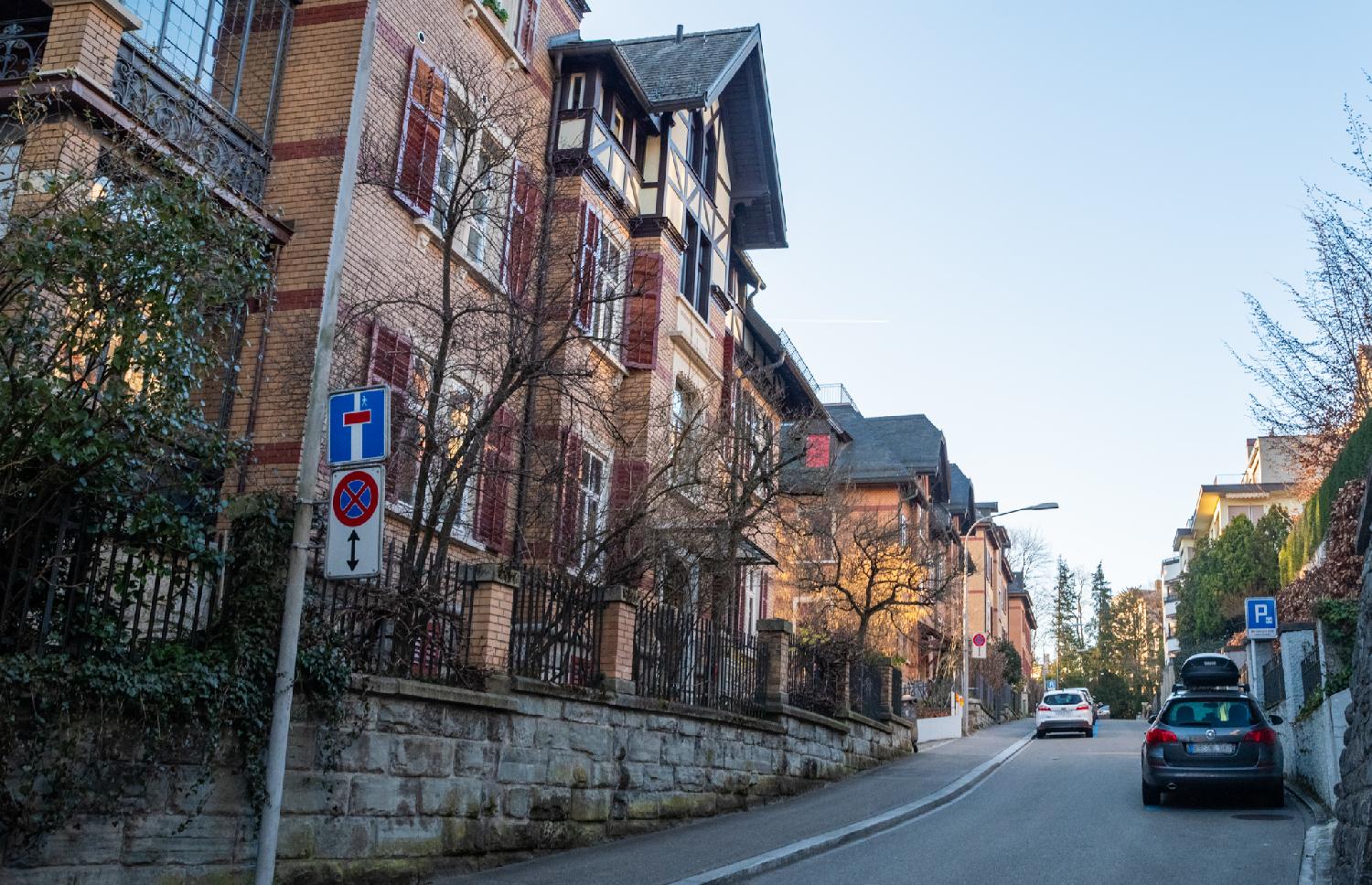 A residential street with imposing buildings with brick façades behind stone and metallic fences and a few cars. The street is marked as a dead-end except for pedestrians.