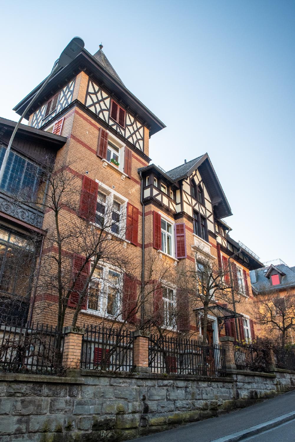 A 4-story residential brick building with dark red blinds, with higher elements covered in timber-frame structures. The building looks imposing and is behind a stone metallic fence and deciduous trees with no leaves.