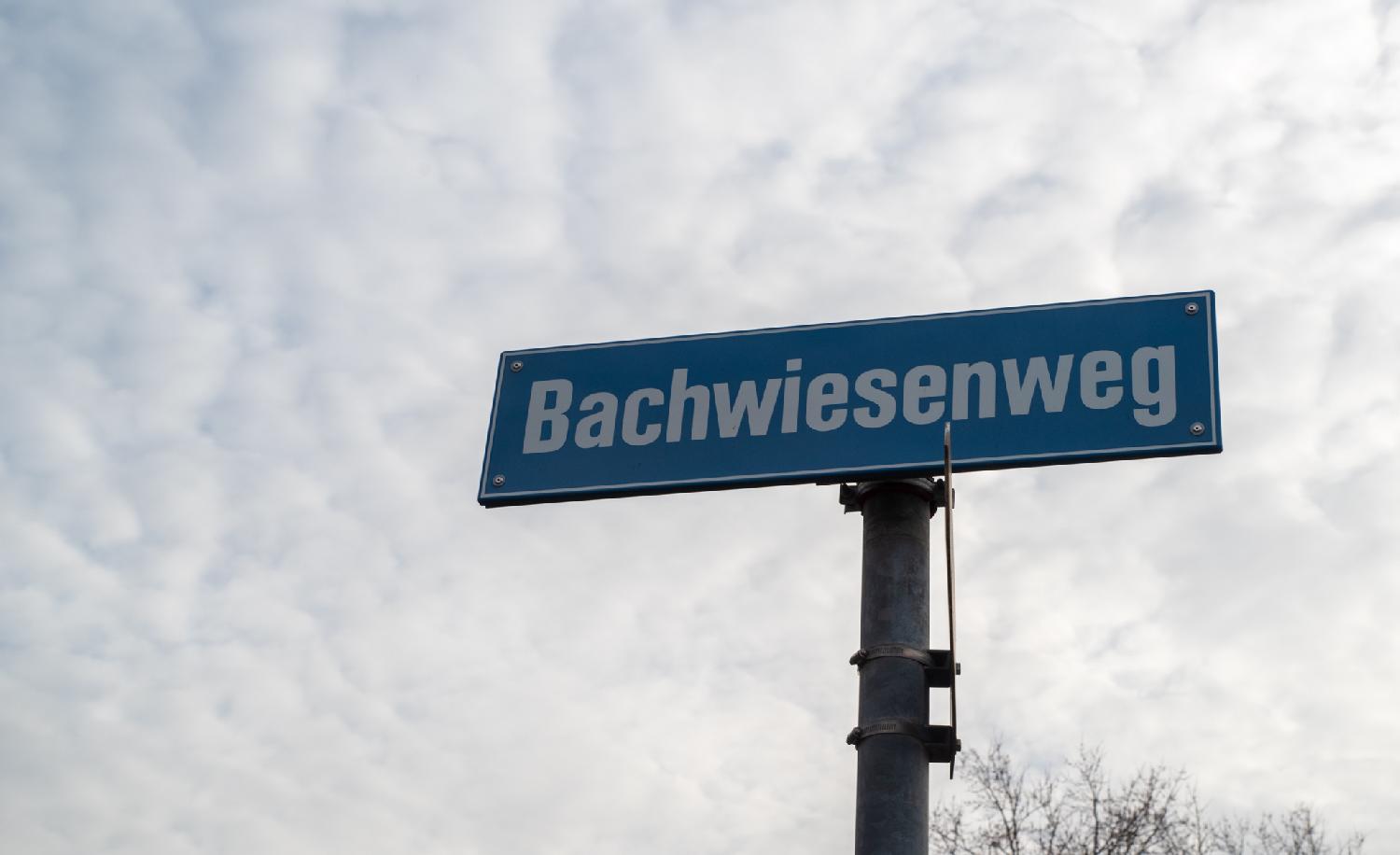 A metallic pole with a blue street sign for Bachwiesenweg, in front of fluffy-looking clouds.