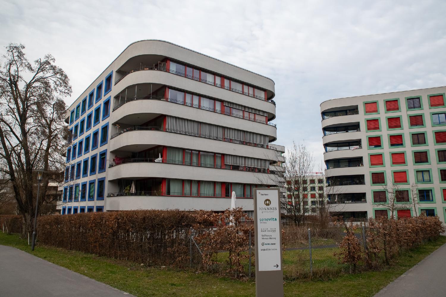 Residential buildings, 6-story high with curved angles. The windows have colorful frames (blue one one, water green on another) and have red/orange blinds; the windows indicate dense habitats, maybe small studios. Two sides of each buildings have longer windows and balconies.