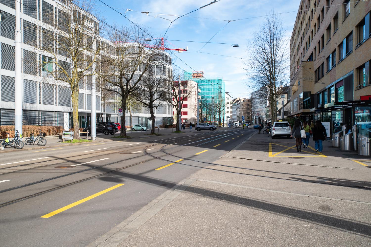 A wide street between 5-6 stories buildings, some tram rail tracks, a few trees and a tram in the background.