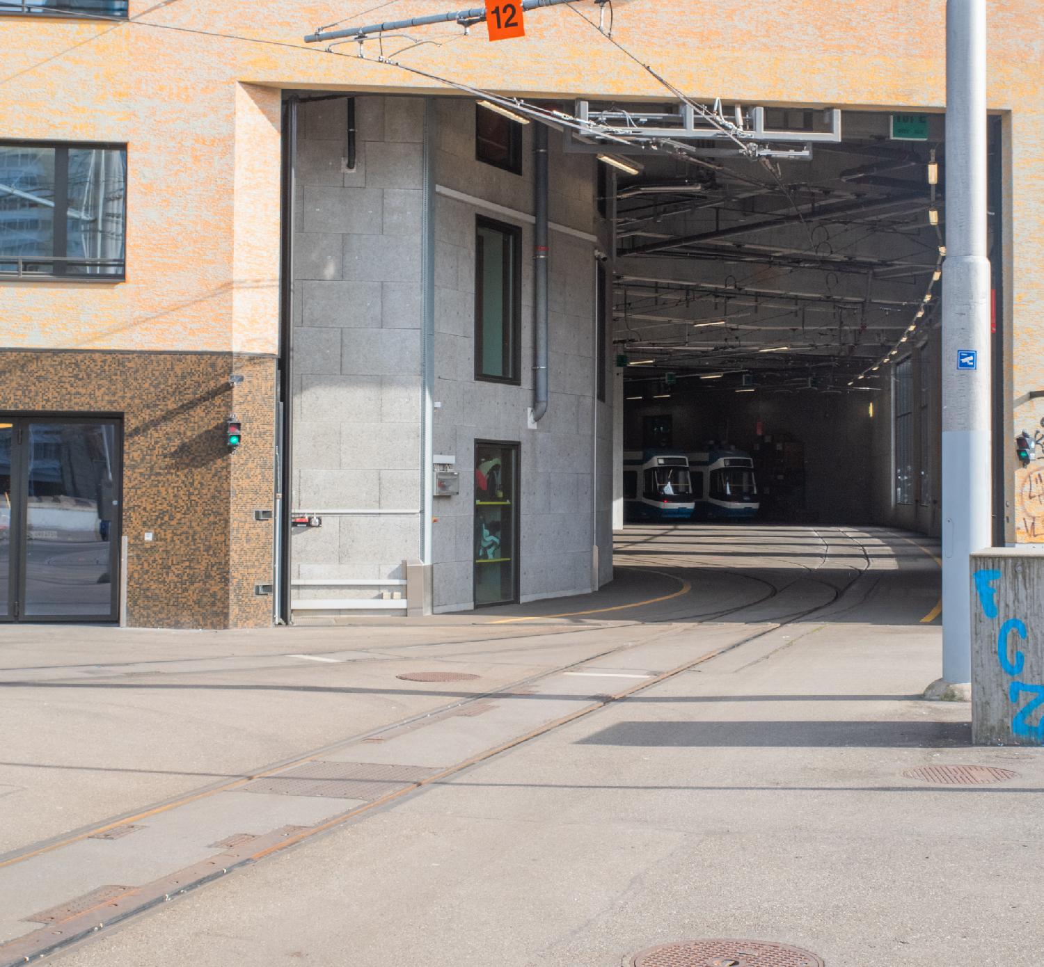 The entrance of a tram depot, with tram tracks leading to it, and a couple of trams visible inside the depot.