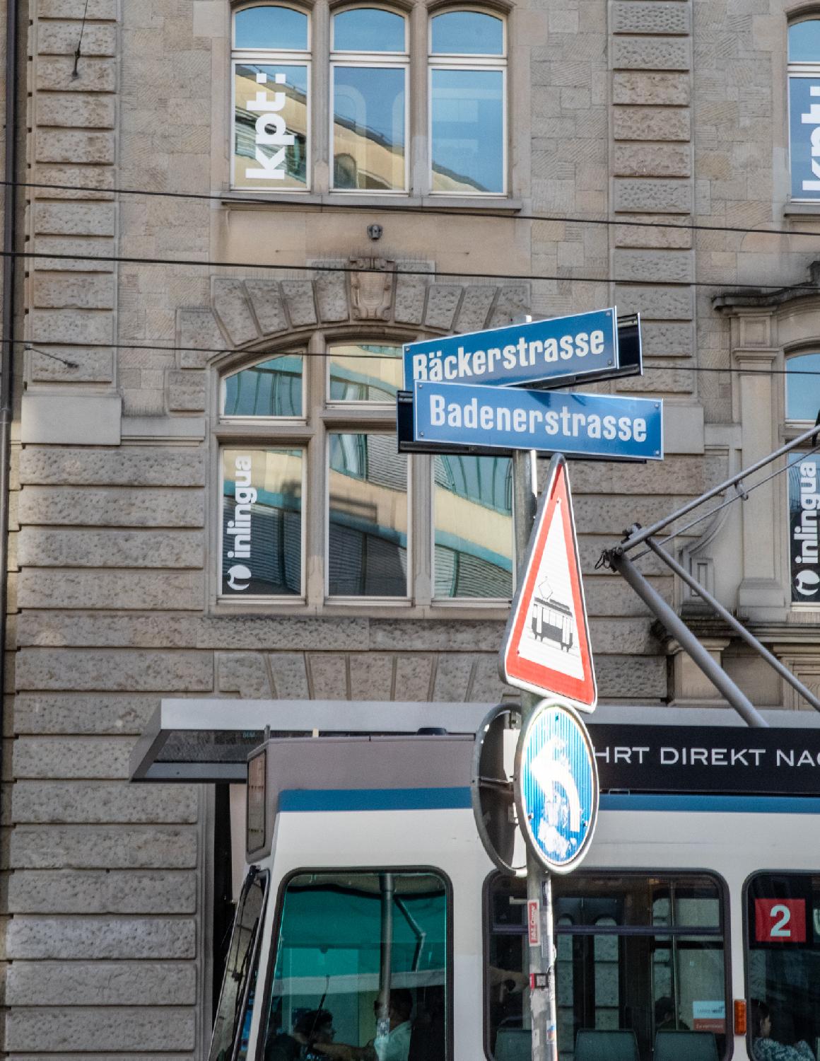 Blue street signs for Bäckerstrasse and Badenerstrasse, on a metallic pole. The background shows a building and the top of a tram is visible between the signs and the building.