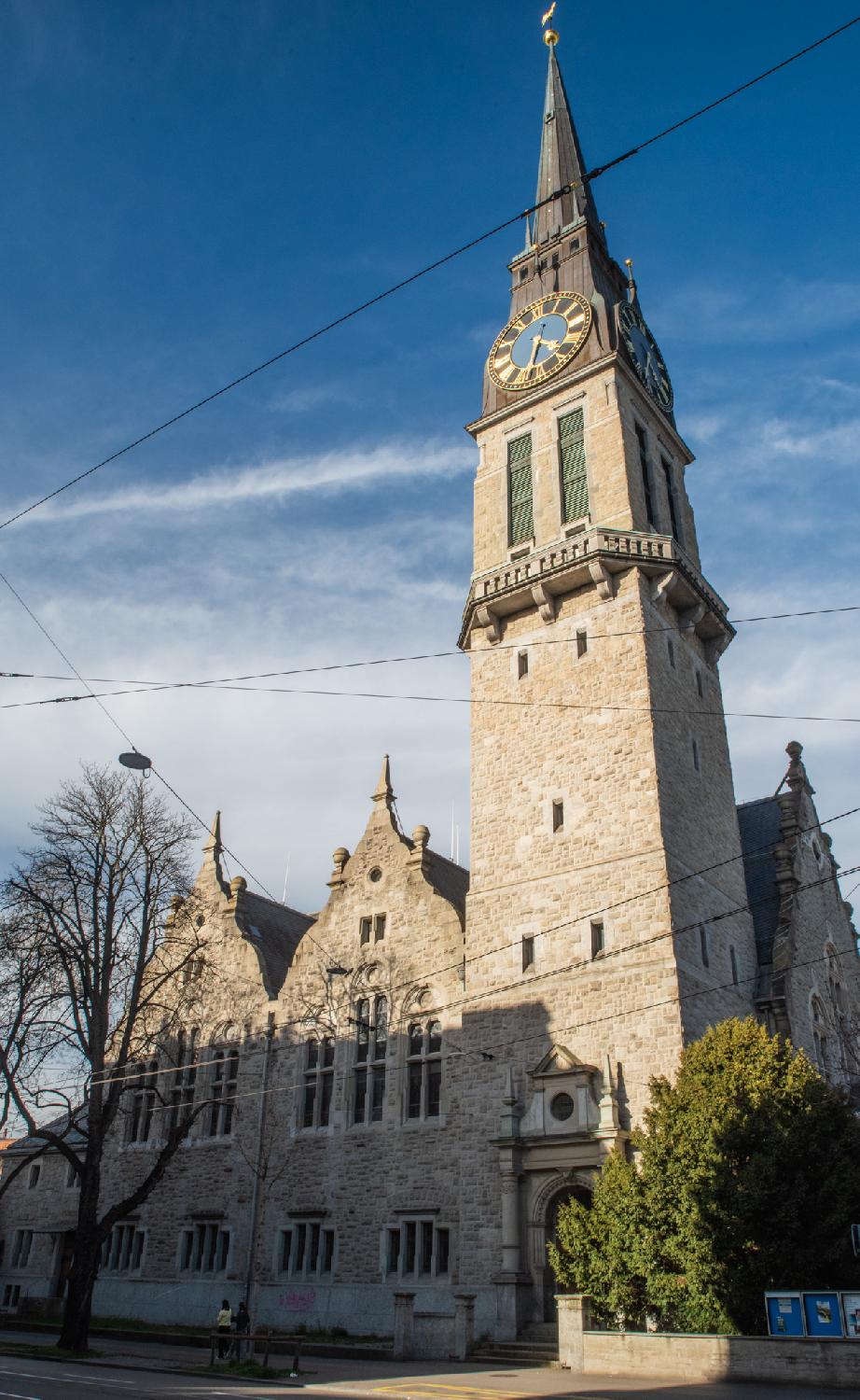 Side of a church with a church tower and a church building in two parts. The church tower has a blue clock and tiny windows; the two parts of the building are identical and show elongated windows, rounded at the top, and curved gabled roofs.