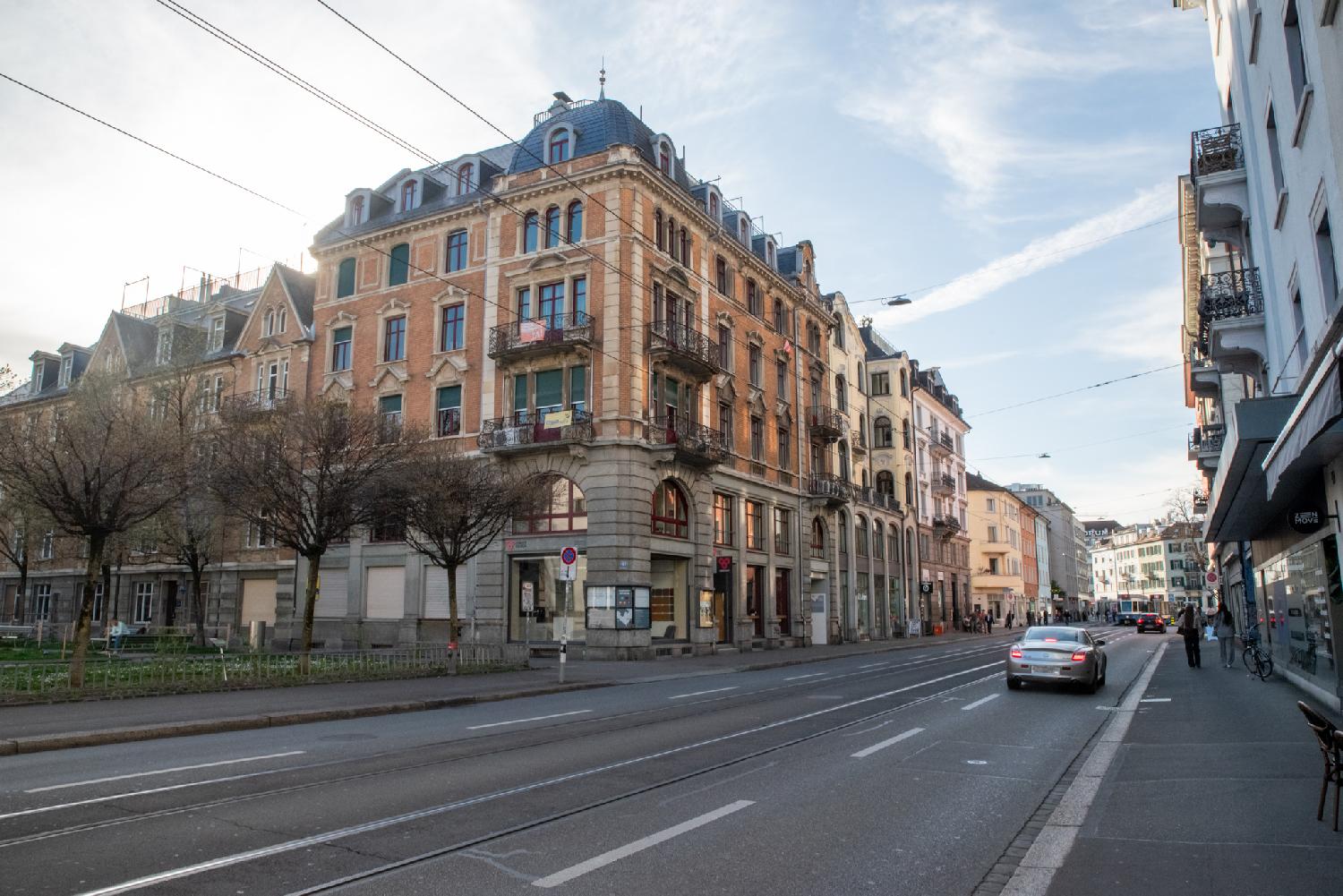 A wide street with tram lines in the middle of two car lanes, between 6-stories buildings. There's a small park on the left side of the street.