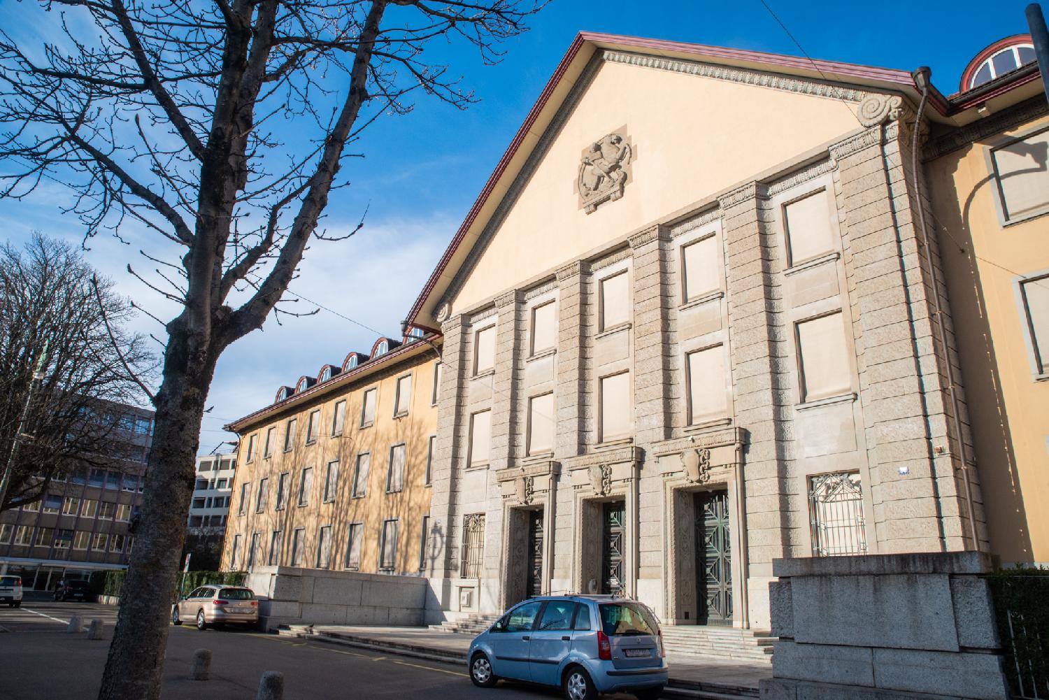 A 4-story official building with a middle part with a gable roof and large metallic gates and two wings with rounded dormer windows.