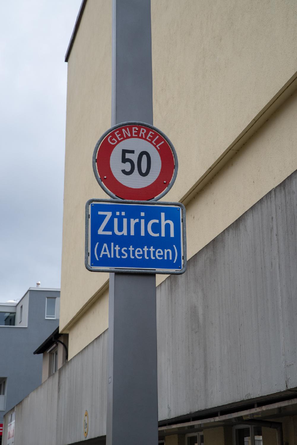 A grey pole with a blue "Zürich (Altstetten)" sign and a 50 speed limit sign in front of non-descript buildings.
