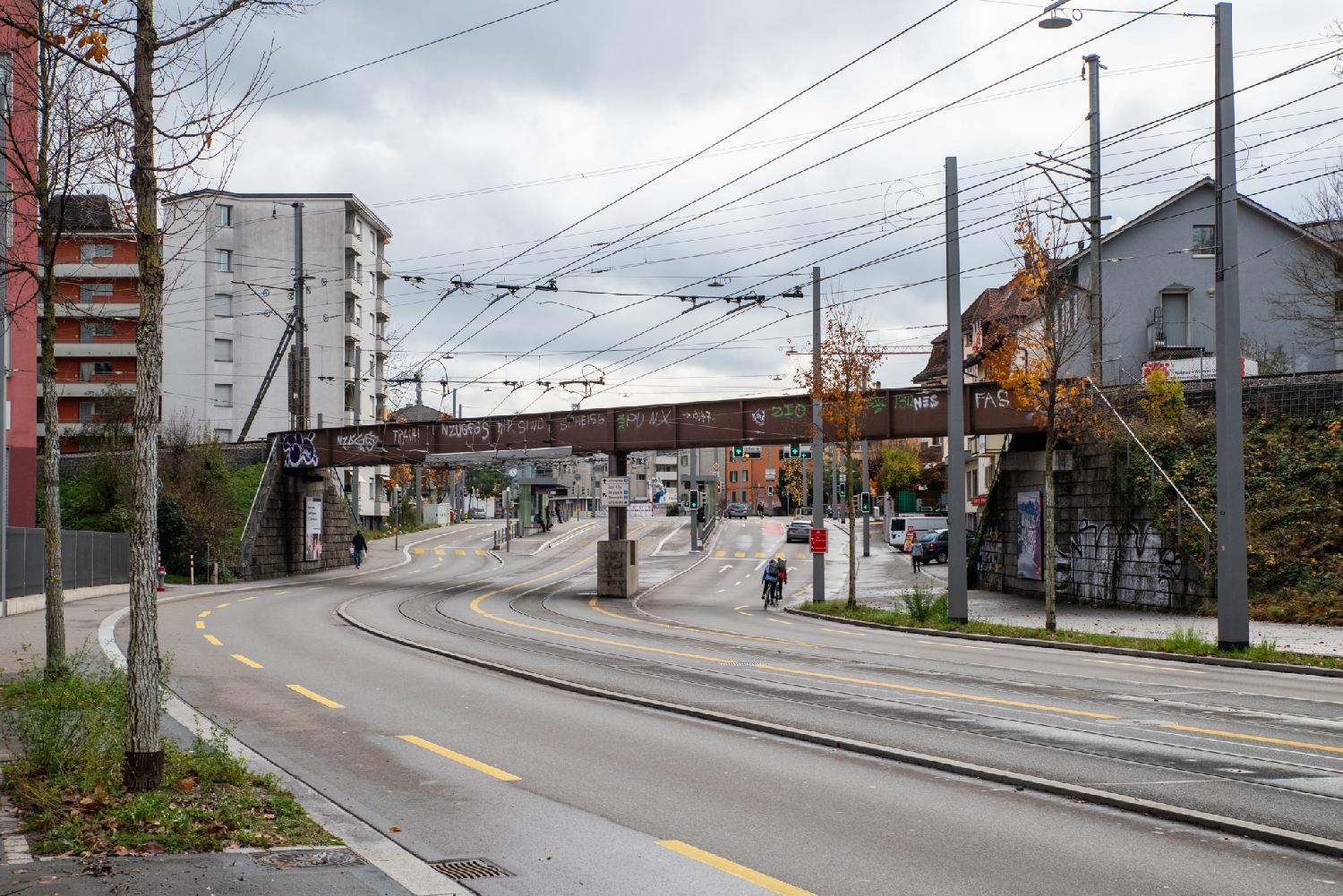 A wide road with two tram tracks in the middle and a train bridge on top, between 3-to-7-story buildings. A tram stop is visible in the background.