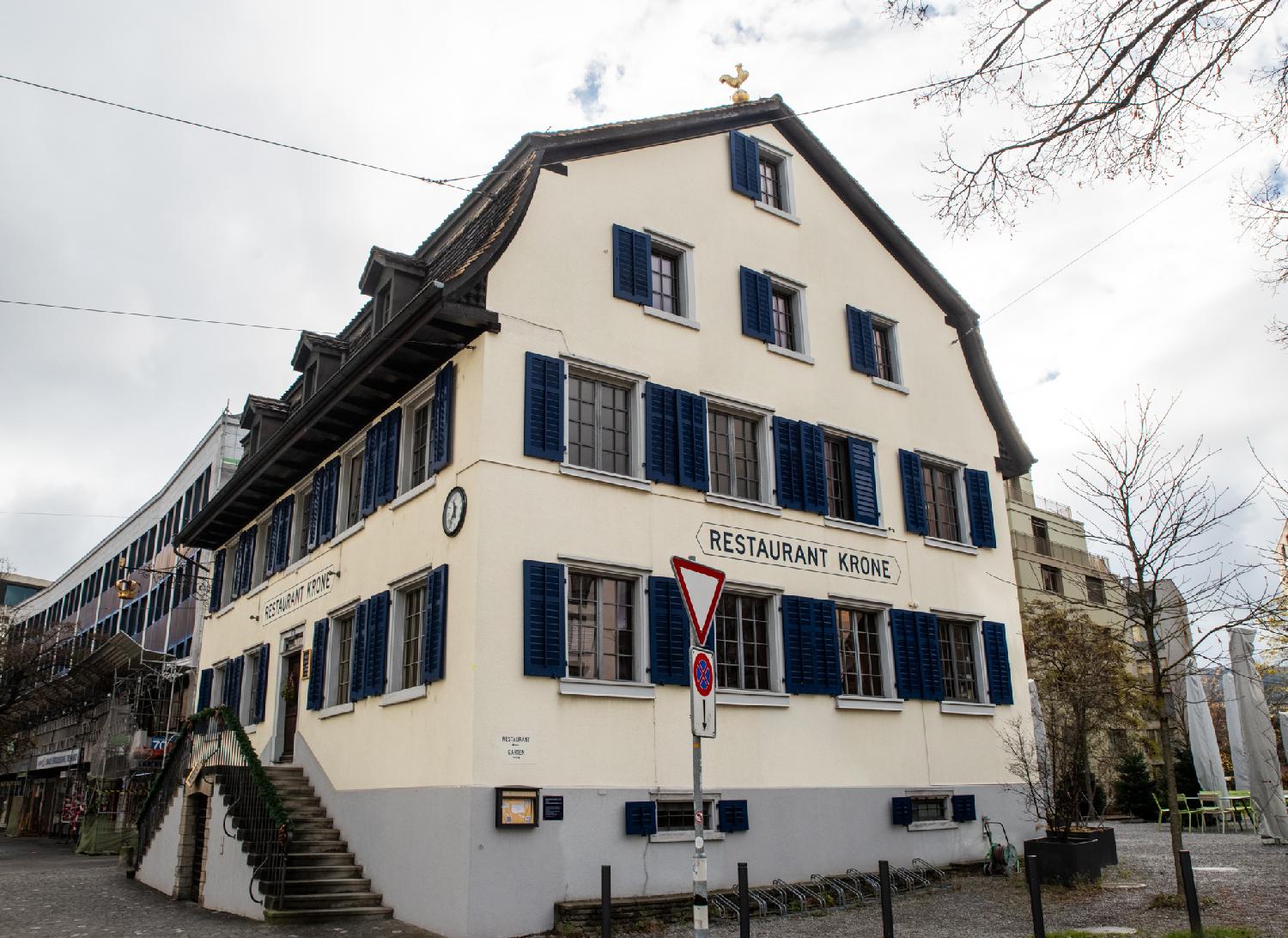 A 4-story with a gambrel roof, seen from the corner, with windows with dark blue blinds. The entrance is on top of stairs and the name Restaurant Krone is painted on the two visible facades.