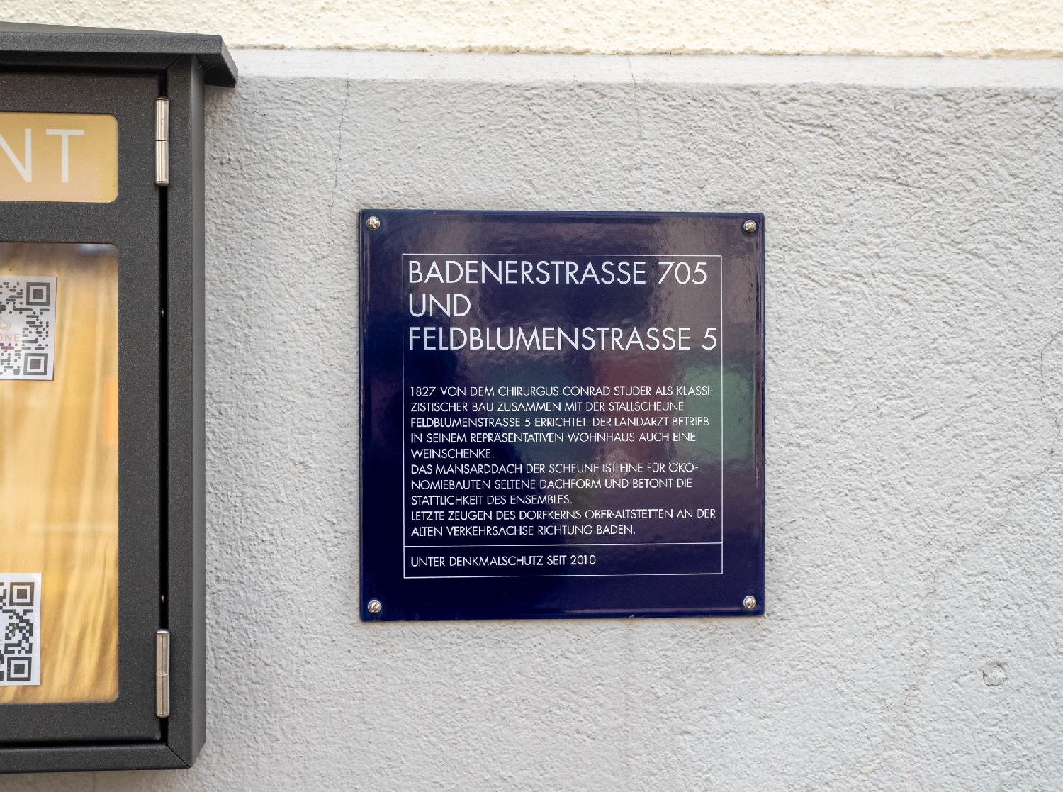 A blue plate on a wall reading "Badenerstrasse 705 und Feldblumenstrasse 5 - 1827 von dem Chirurgus Conrad Studer as classizistischer Bau zusammen wit der Stallscheune Feldblumenstrasse 5 errichtet. Der Landartzt betrieb in seinem repräsentativen Wohnhaus auch eine Weinschenke. Das Mansarddach der Scheune ist eine für Ökonomiebauten seltene Dachform und betont die Stattlichkeit des Ensembles. Letzte Zeugen des Dorfkerns Ober-Altstetten an der alten Verkehrsachse richtung Baden. Unter Denkmalschutz seit 2010."