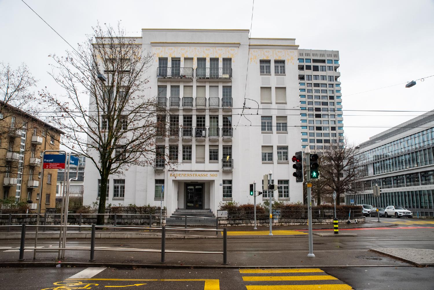 A white imposing 5-story office building, with a middle part with clustered windows and two wings. There is some decorative yellow/gold floral paintings on top of the building. The address of the building (Badenerstrasse 580) is painted above the door in golden capital letters. The Kappeli tram stop is visible in the foreground.
