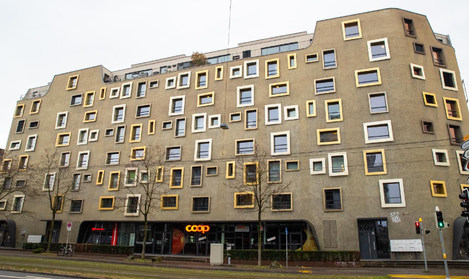 A brown residential building whose windows are not aligned and all framed in different shades of white and yellow. There's a Coop shop on the ground floor.