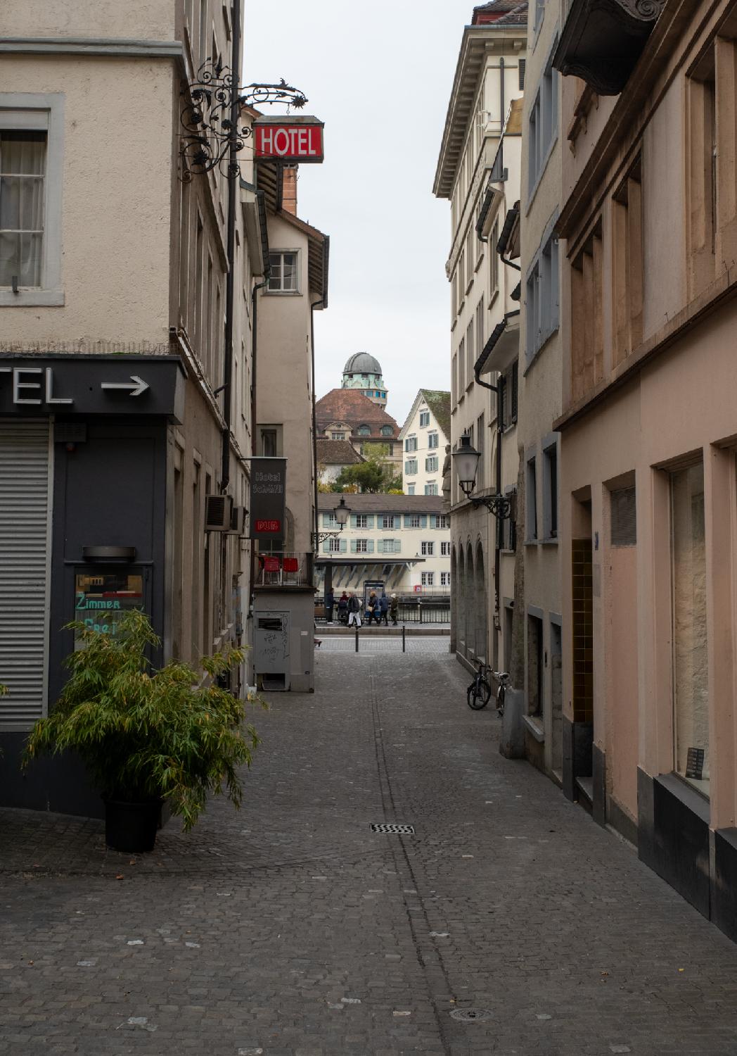 A narrow paved street with 5-6-story buildings on each side. A red HOTEL sign is protruding from the left wall. In the background, a tower with a round observatory roof, and the river flowing perpendicularly in front of a tram stop.