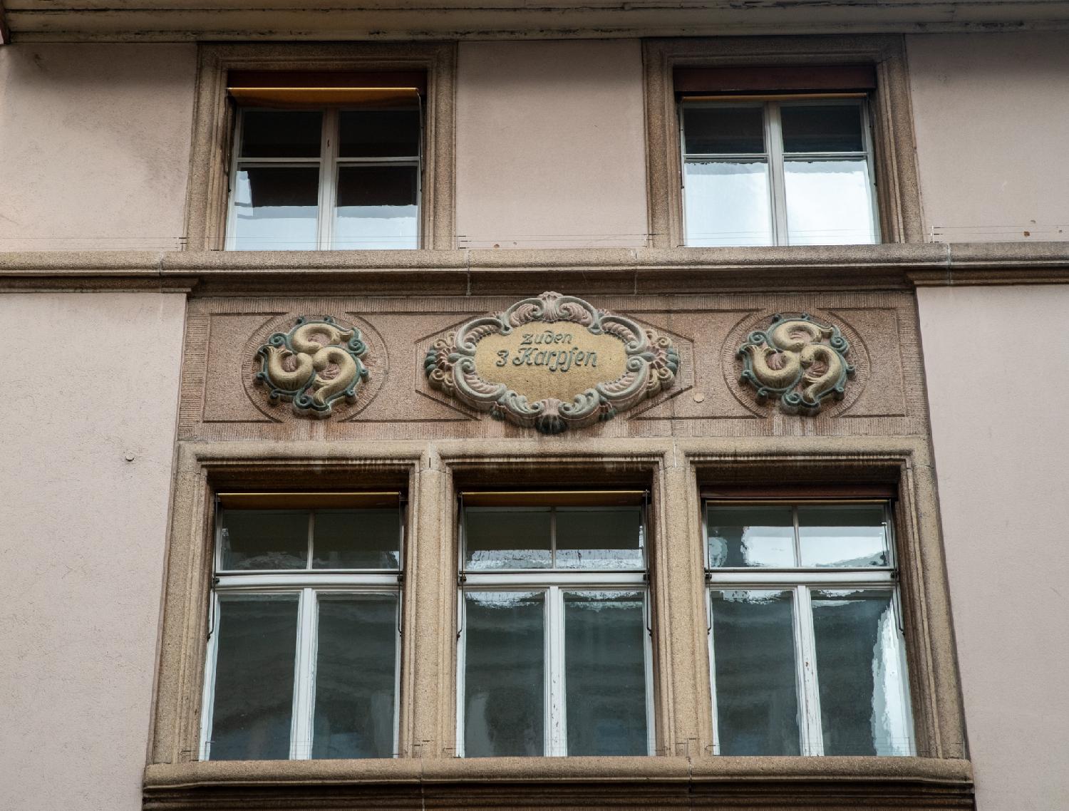 A wall with two rows of windows separated by a relief. The top row has two windows, the bottom row has three windows. The relief has a center inscription "zuden 3 Karpfen" (the 3 carps) with, on each side, a yellow and green medallion with three carps meeting in the center.