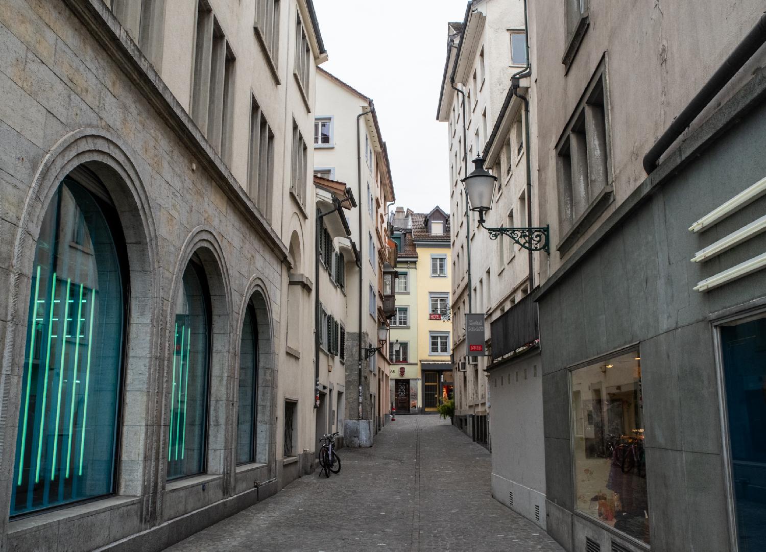 A narrow paved street between buildings 3 to 6 storey high. The building on the front left has arched windows with vertical green neon stripes in them.