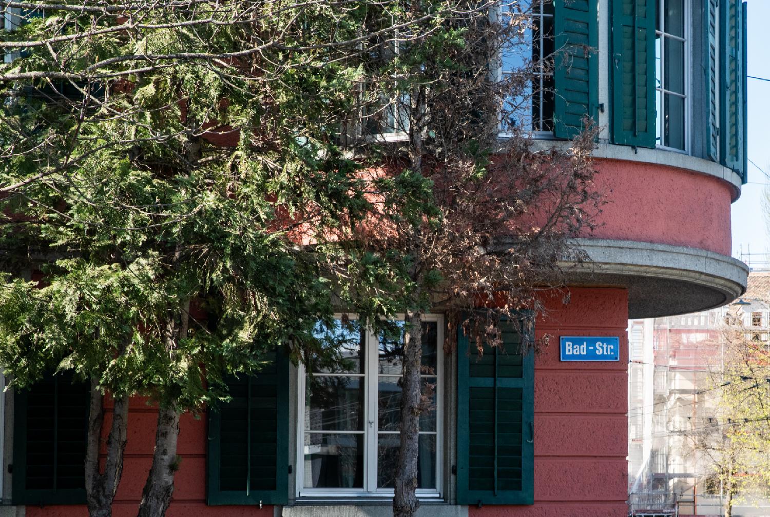 A crop of a red residential building with round bay windows and dark green blinds, with a blue street sign showing "Bad-Str." The building is partly hidden by a tree.