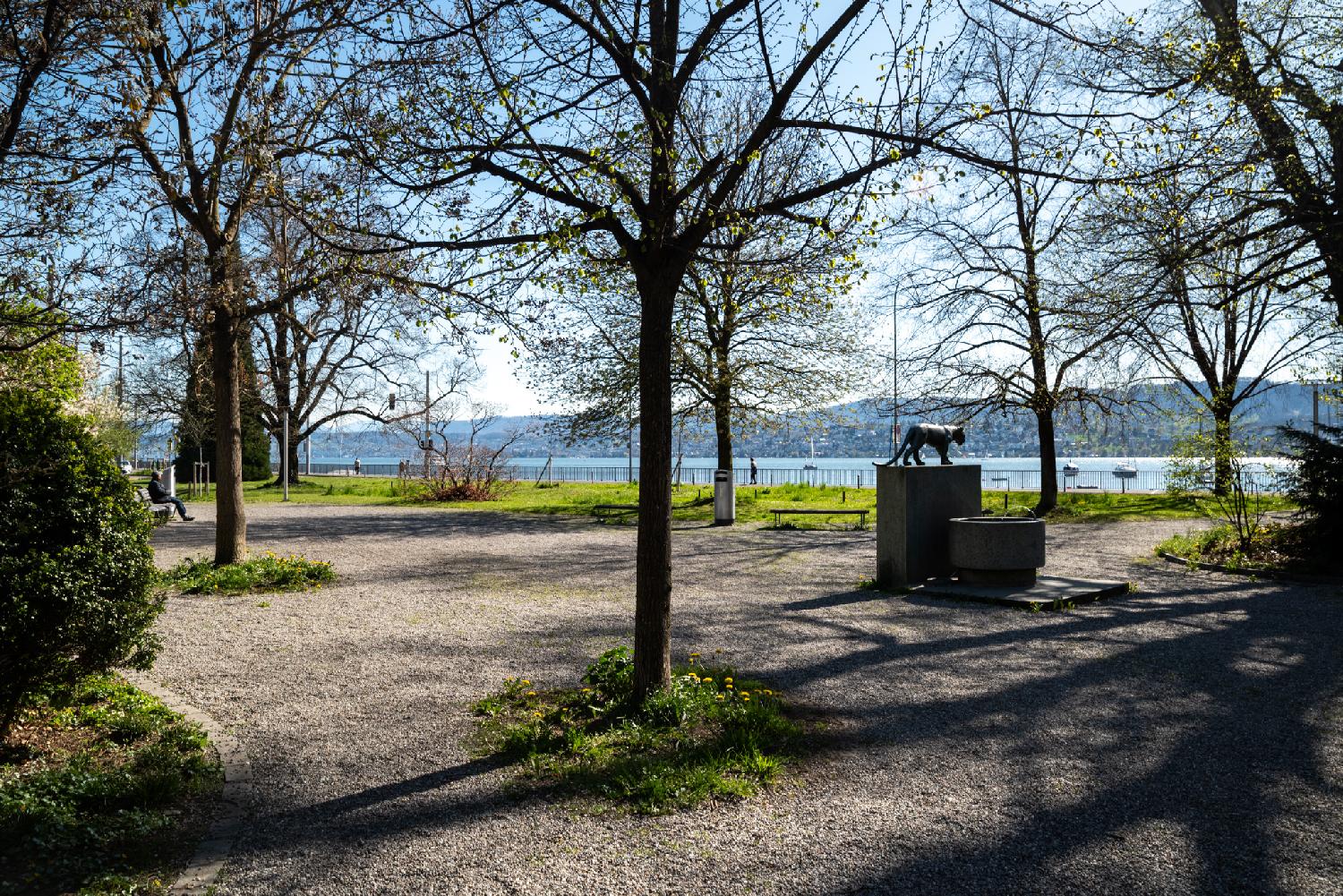 A small park next to a lake with a few trees, some benches and a drinking fountain topped by a panther sculpture.